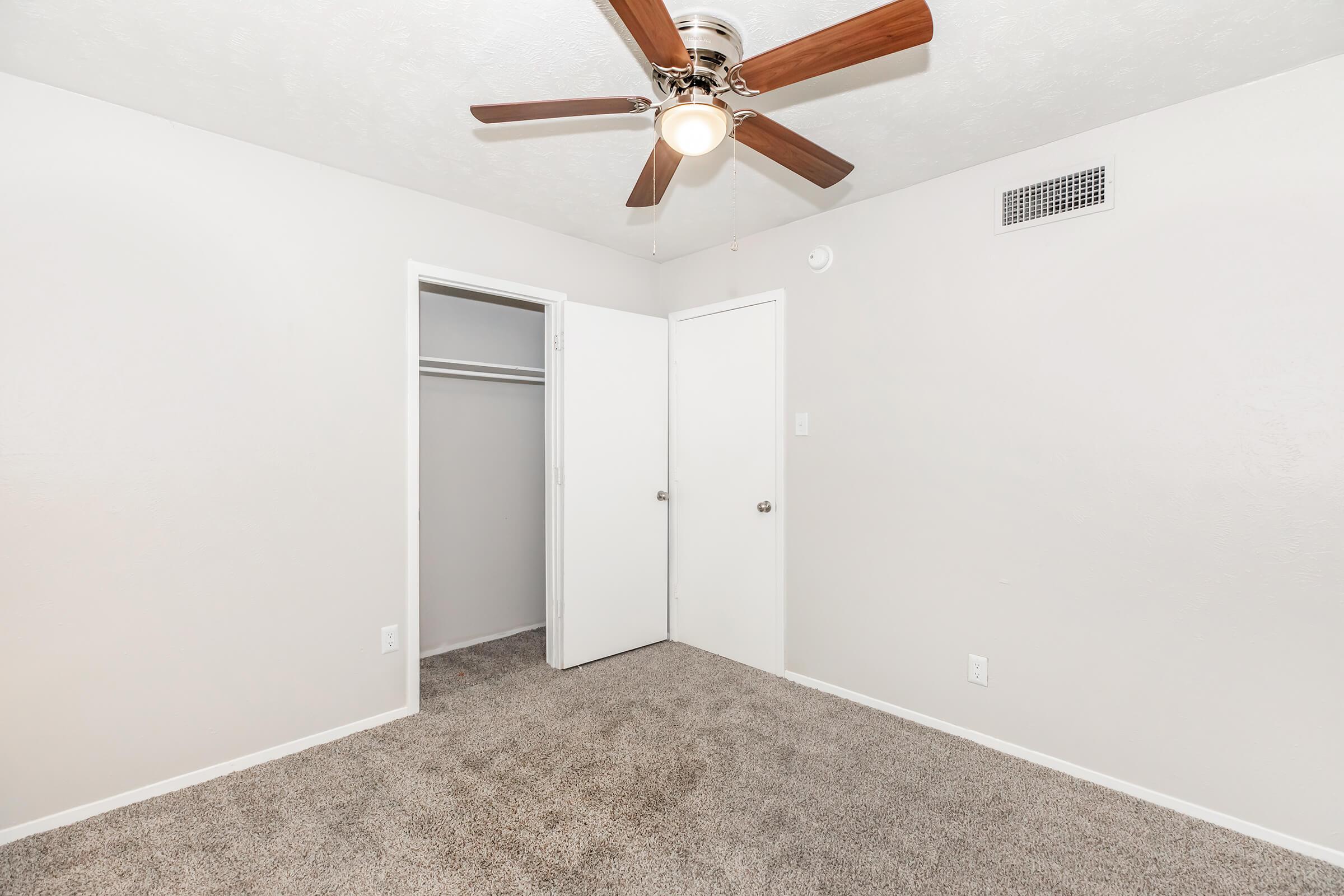 A light-colored room featuring a ceiling fan, beige carpeting, and two doors—one leading to a closet and the other to another space. The walls are painted a soft gray, creating a bright and airy feel.