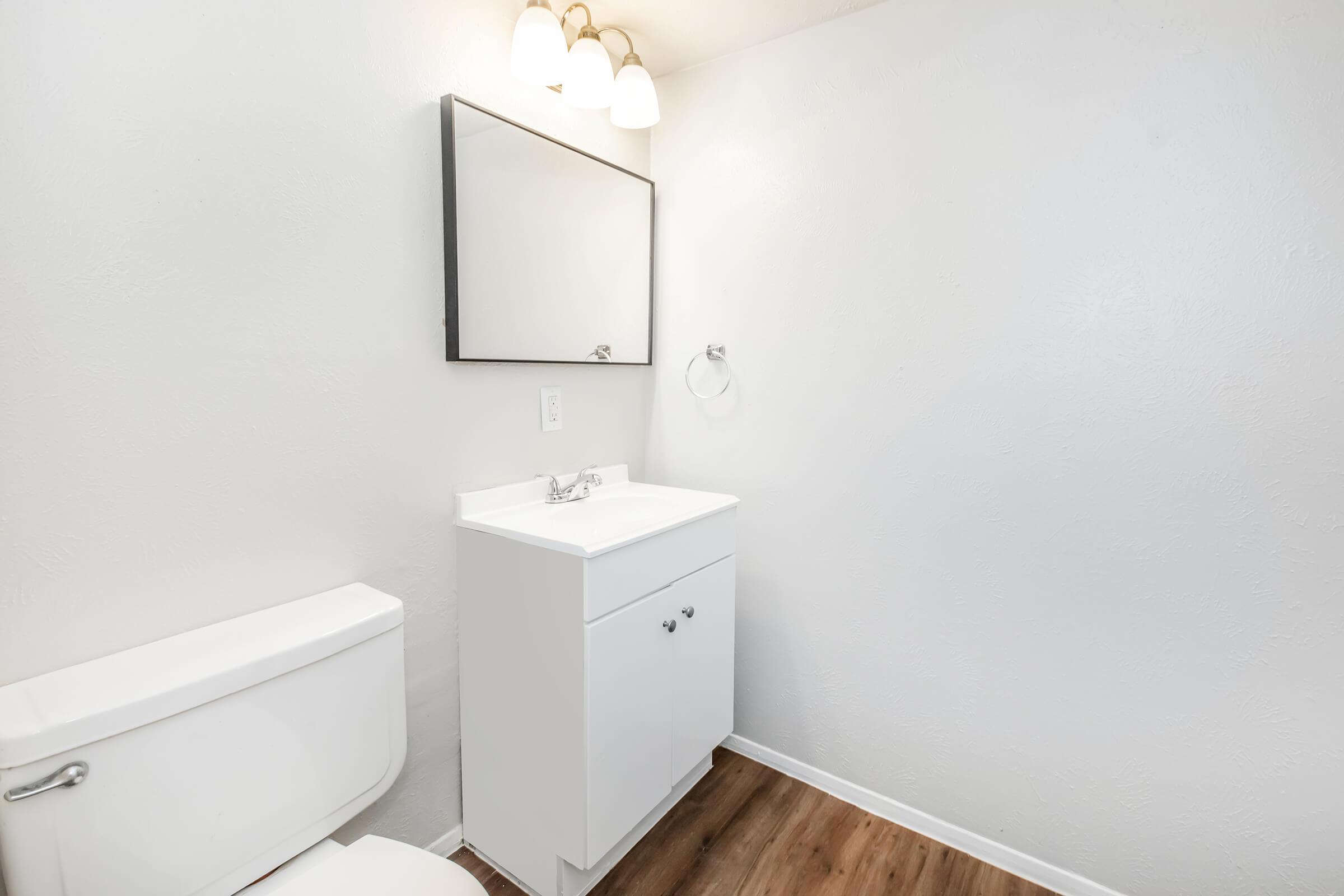 A clean, modern bathroom featuring a white toilet, a small white vanity with a sink, and a rectangular mirror above the vanity. The walls are painted in light colors, and there is a towel ring on the wall. Soft lighting is provided by three ceiling-mounted light fixtures. The floor is a warm wood tone.