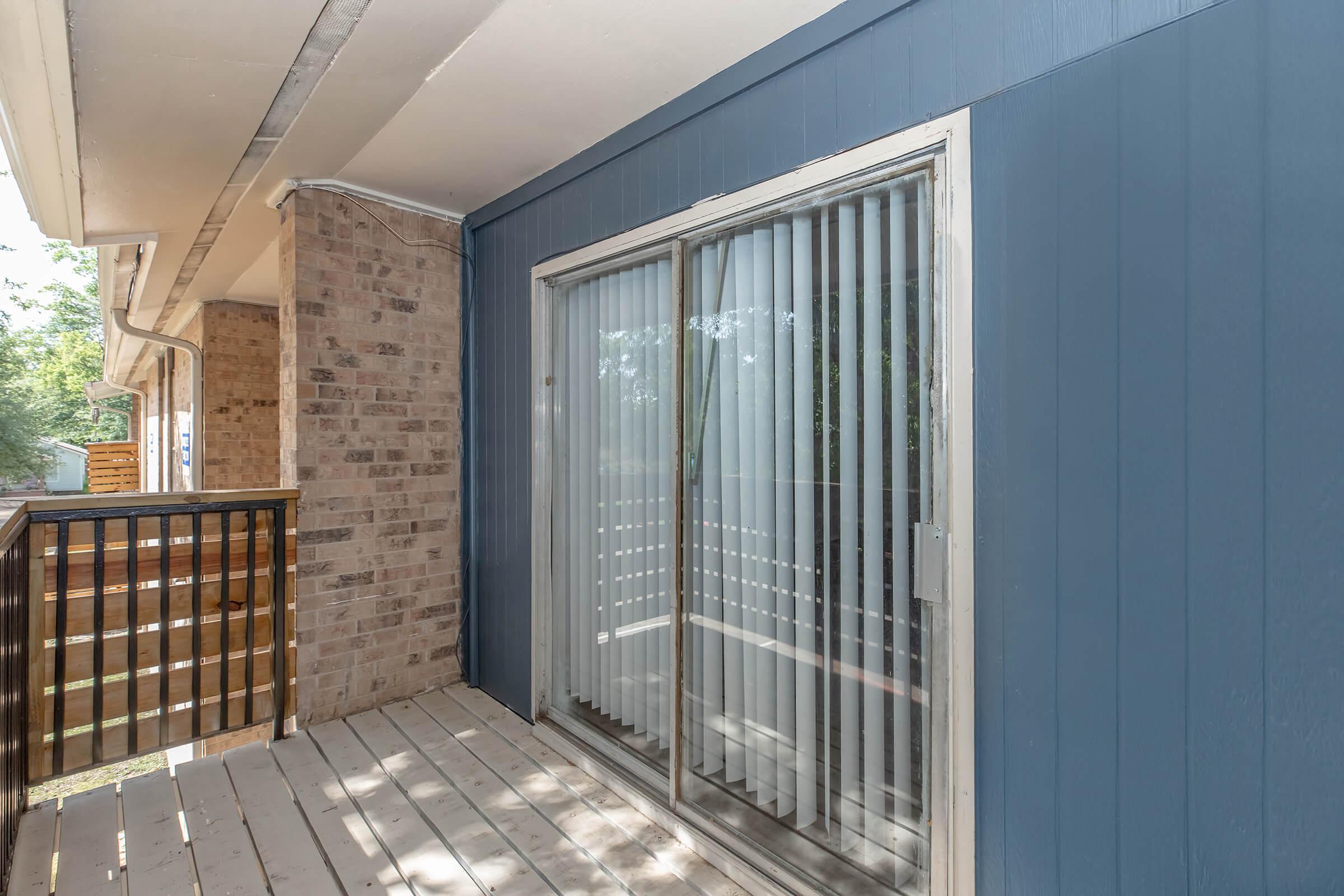 A view of a small balcony featuring a sliding glass door with vertical blinds. The exterior wall is blue, and there's a wooden railing on one side. The floor is made of wooden planks, and some greenery is visible outside the balcony, indicating a natural environment.
