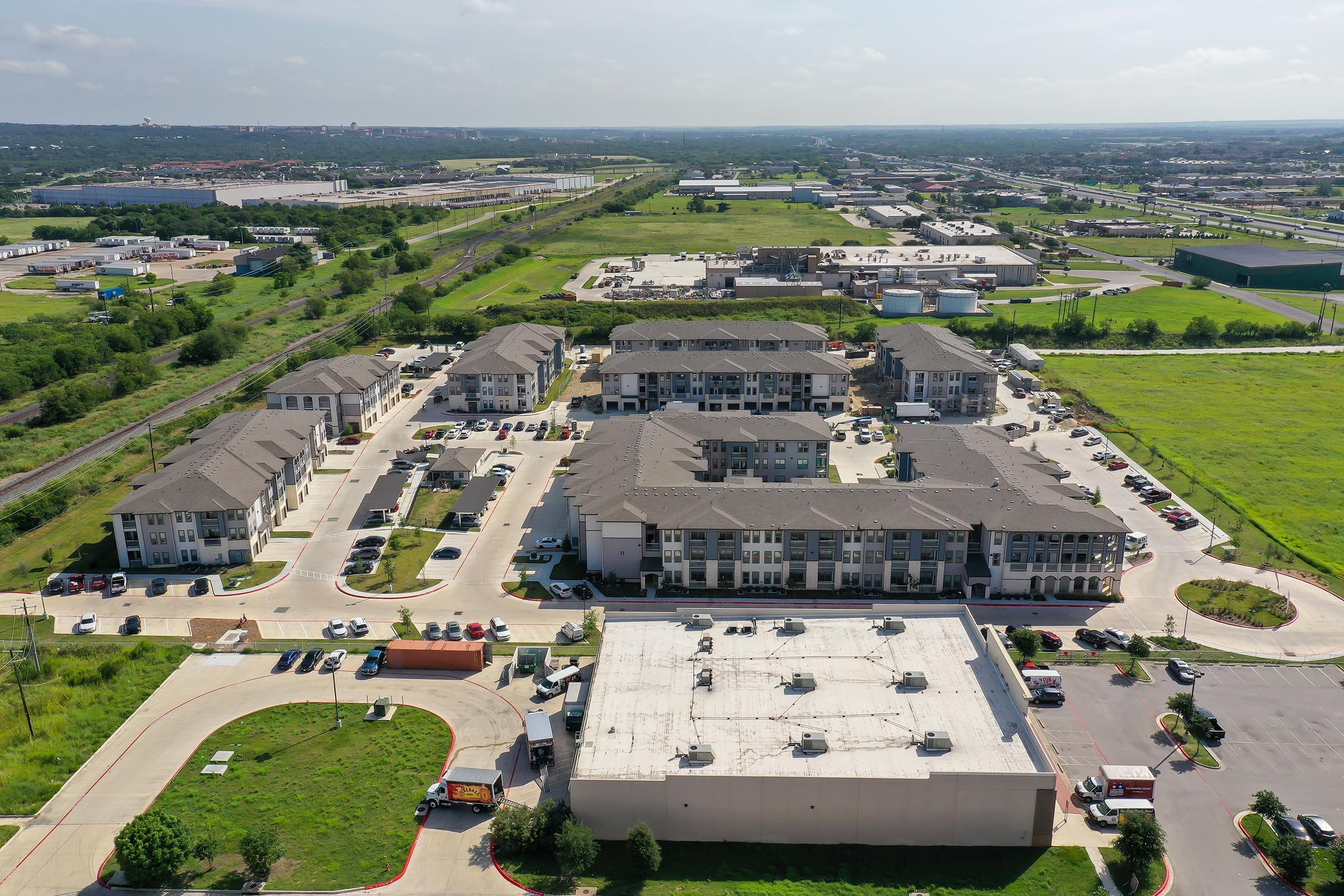 Aerial view of a residential area featuring multiple apartment buildings with surrounding parking lots. Green fields and commercial buildings are visible in the background, along with infrastructure such as roads and tracts of land. The scene depicts a mix of urban development and natural landscape.