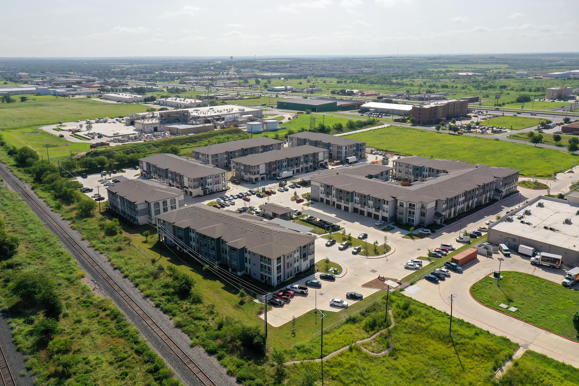 Aerial view of a suburban apartment complex surrounded by grassy areas and industrial buildings. The complex features several multi-story buildings with parking lots in front. In the background, a railway line runs next to the properties and larger commercial buildings are visible farther away.