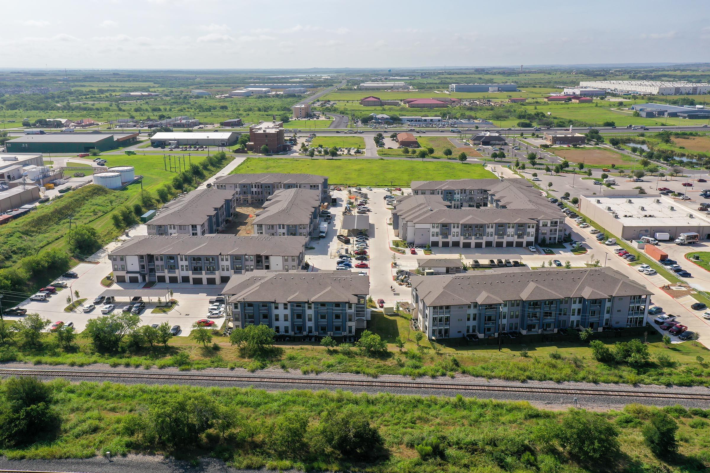 Aerial view of a residential apartment complex surrounded by greenery and open fields. The area includes multiple buildings organized in a cluster with parking lots. In the background, various structures and fields stretch out, indicating a suburban environment with small businesses and developments nearby.