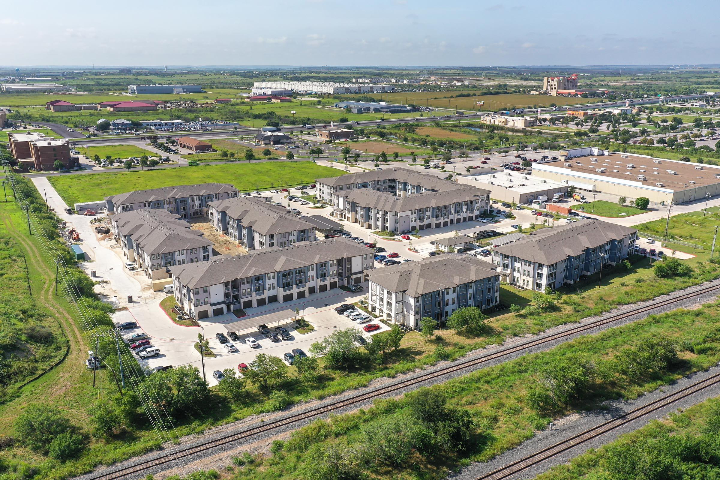 Aerial view of a suburban area featuring several residential apartment buildings surrounded by parking lots and green spaces. In the background, commercial buildings and fields are visible, along with a railroad track running along the bottom of the image. The sky is clear with few clouds.