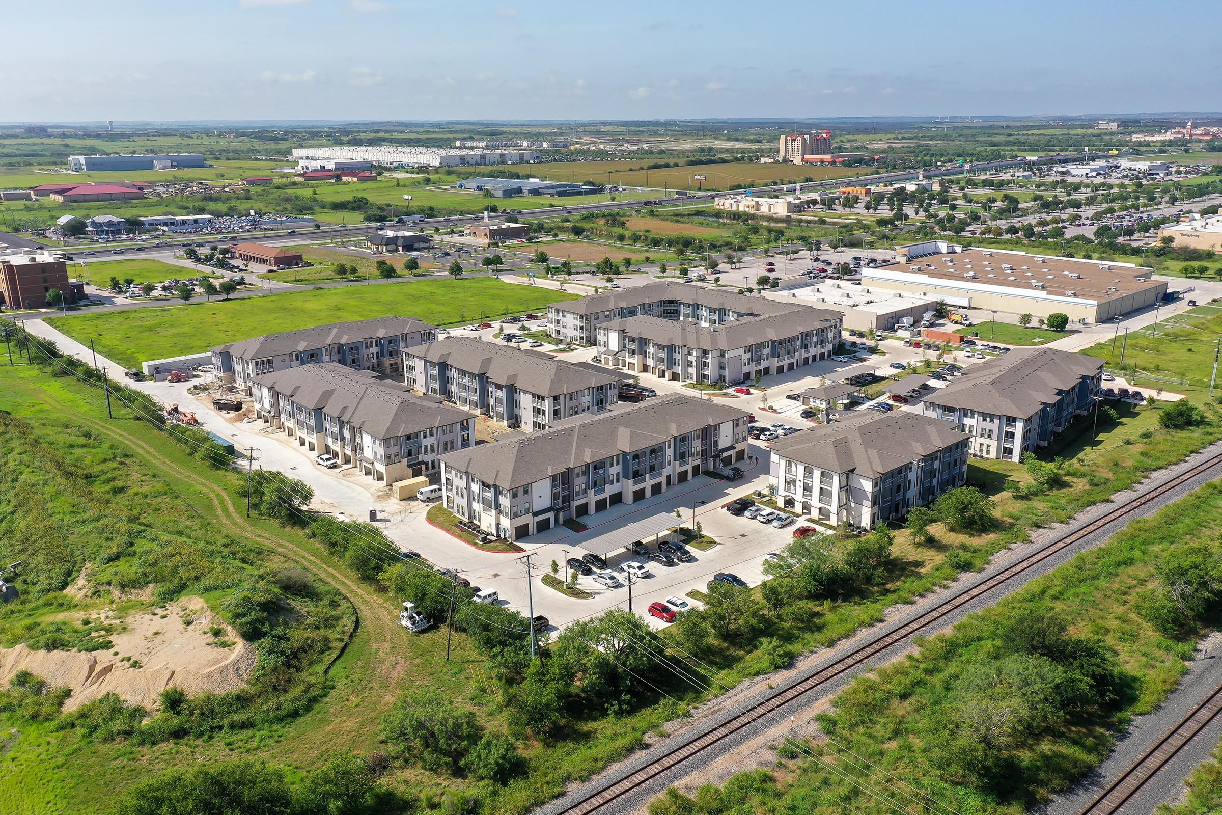 Aerial view of a suburban apartment complex surrounded by green fields and nearby commercial buildings. The complex consists of multiple low-rise buildings arranged around a central parking area, with a railroad track visible in the foreground and various industrial structures in the background.