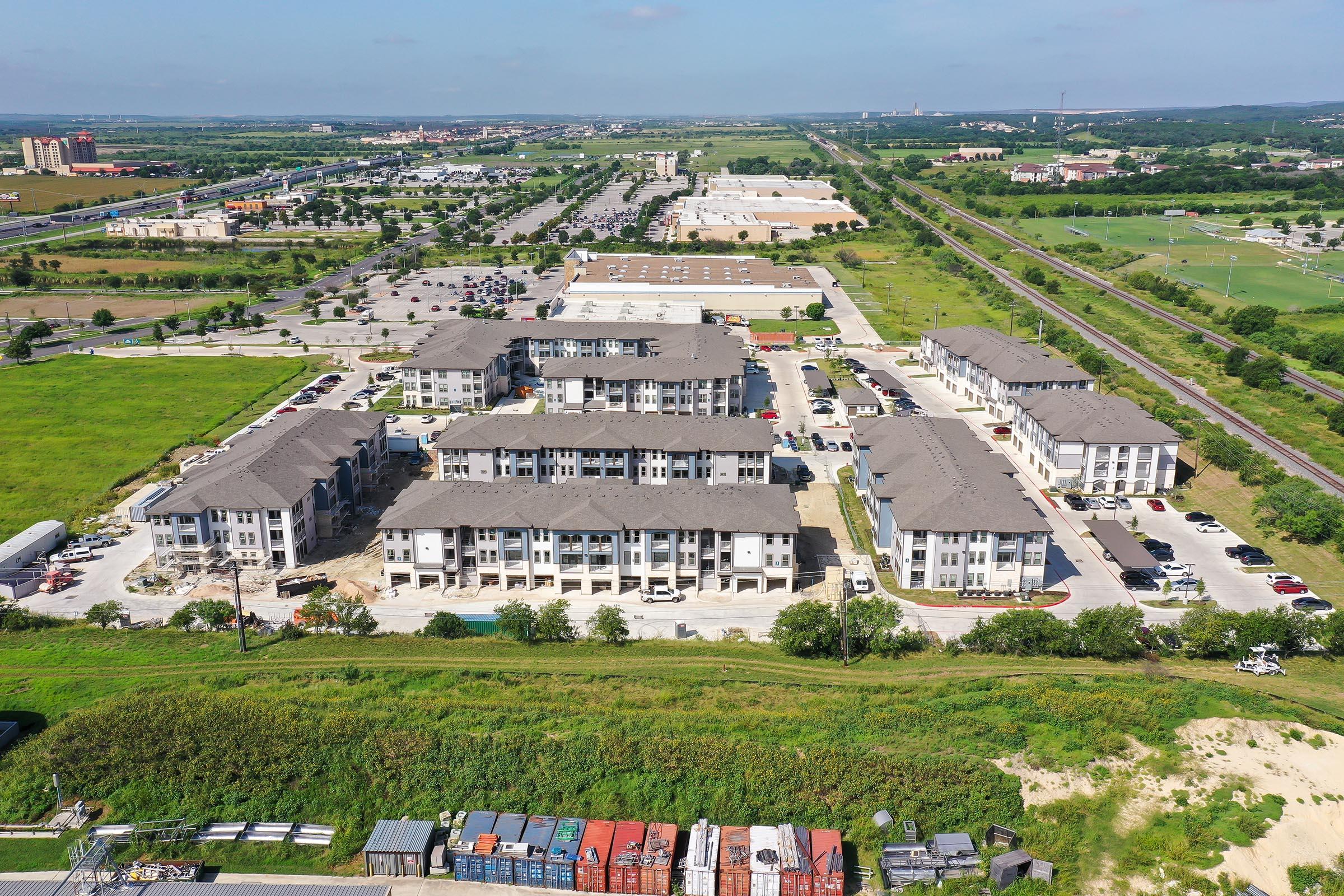 Aerial view of a residential development featuring multiple apartment buildings, surrounded by green fields and parked cars. In the background, commercial areas and roads are visible, highlighting urban growth. The scene showcases a mix of nature and urban living.