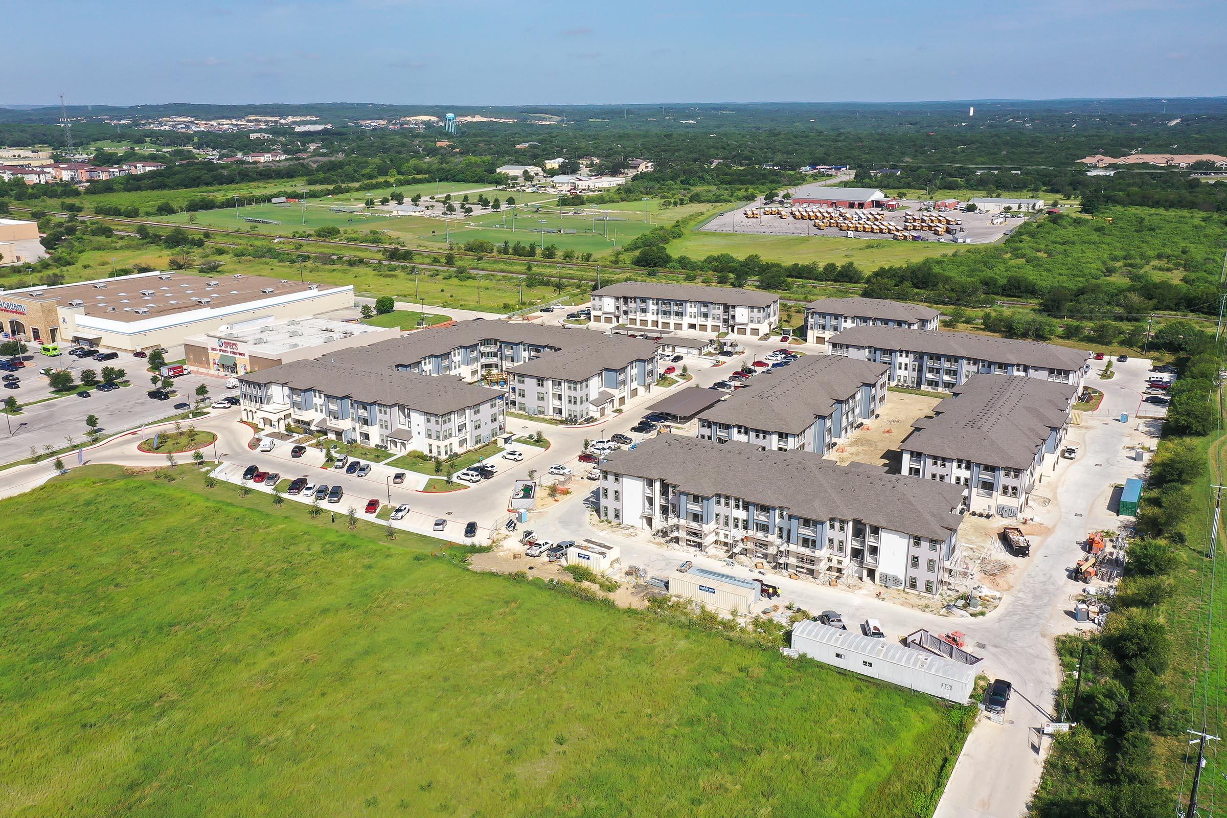 Aerial view of a developing residential area featuring multiple apartment buildings, surrounded by greenery and open land. In the background, there are commercial buildings and parking lots visible, indicating a mixed-use environment. The scene is bright and captures a sunny day.