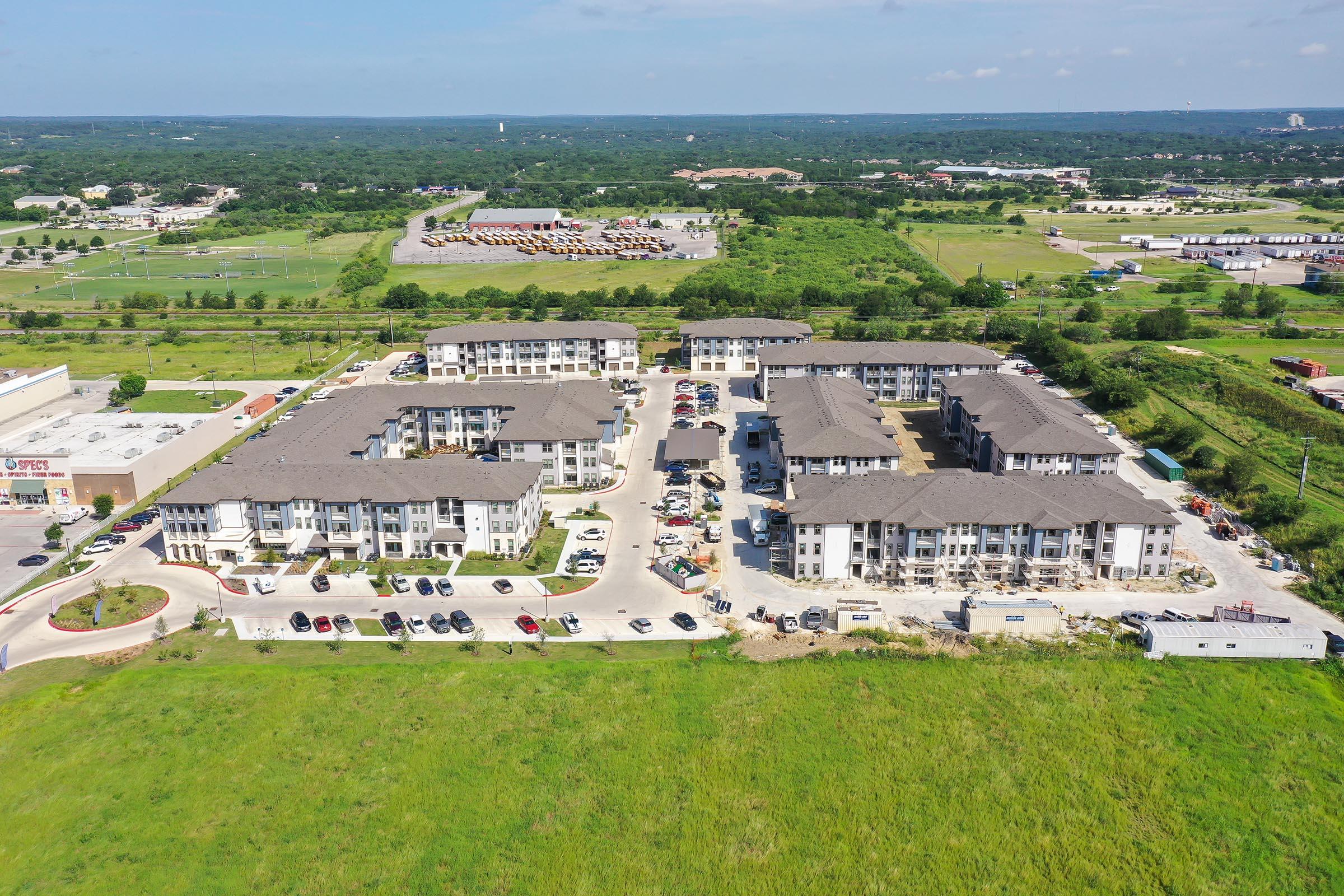 Aerial view of a modern apartment complex with multiple buildings and parked cars, surrounded by greenery and open land, with industrial structures visible in the distance. The scene captures a sunny day with a clear sky and expansive views of the surrounding landscape.