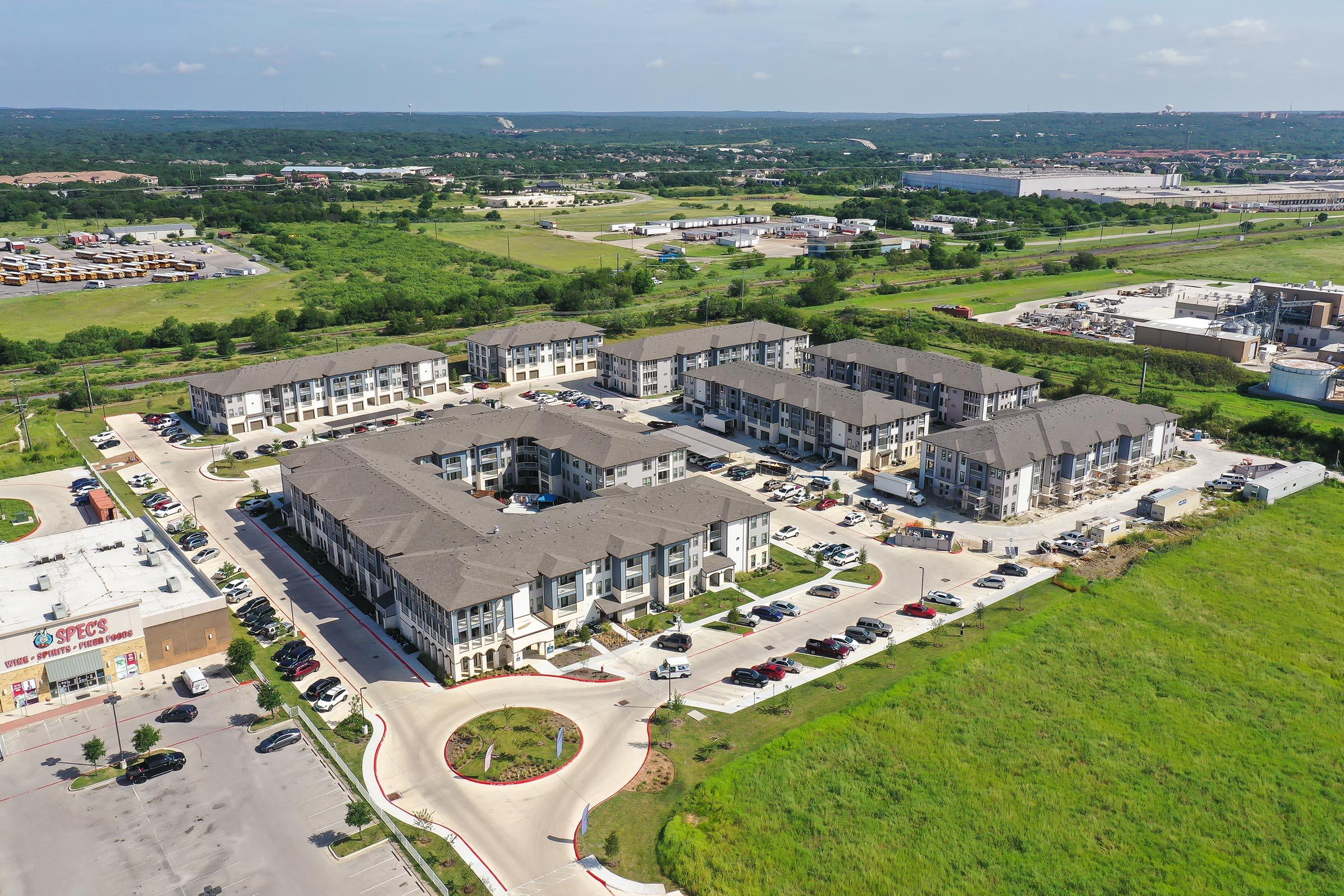 Aerial view of a modern apartment complex with multiple buildings, surrounded by landscaped lawns and parking lots. In the background, commercial buildings and open fields can be seen, indicating a suburban setting with nearby amenities. Clear blue skies above enhance the scene.