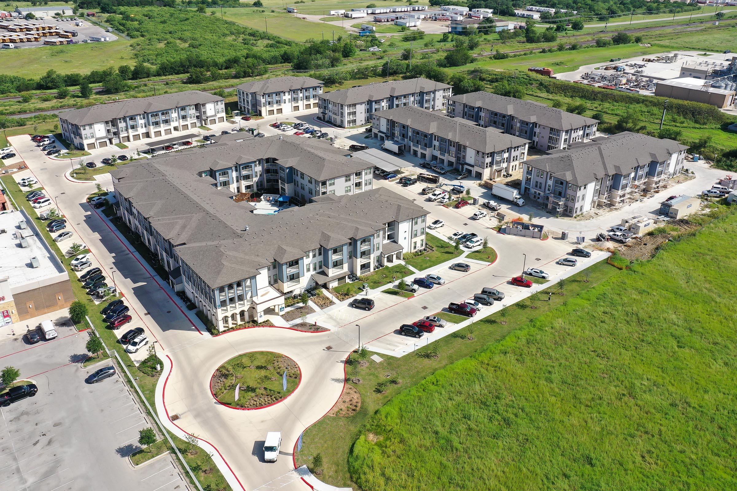 Aerial view of a residential complex featuring multiple apartment buildings arranged in a U-shape, surrounded by parking lots. The area includes landscaped green spaces, with a mix of vehicles parked. In the background, there are commercial buildings and open land, indicating a suburban setting.