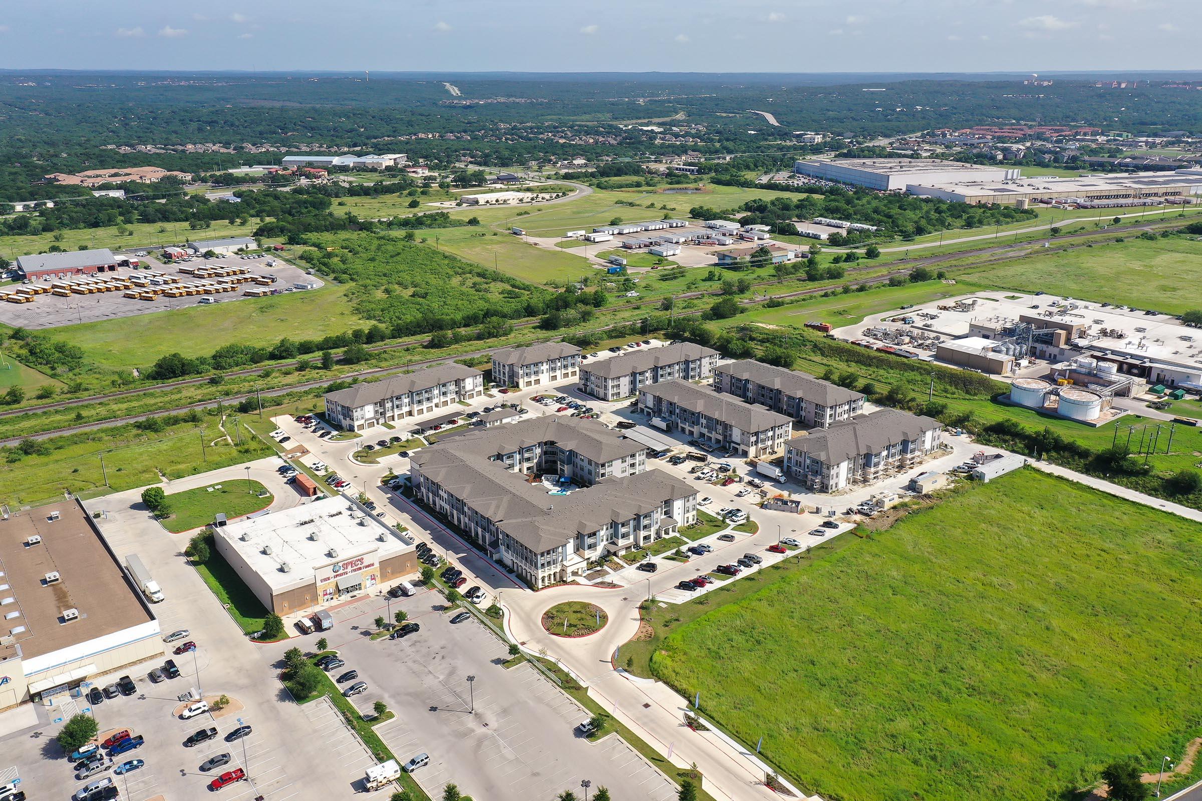 Aerial view of a residential complex surrounded by greenery and open land. The area features several buildings with a parking lot, nearby commercial spaces, and roads. In the background, there's a train line and additional commercial development, showcasing a mix of urban and rural landscapes.