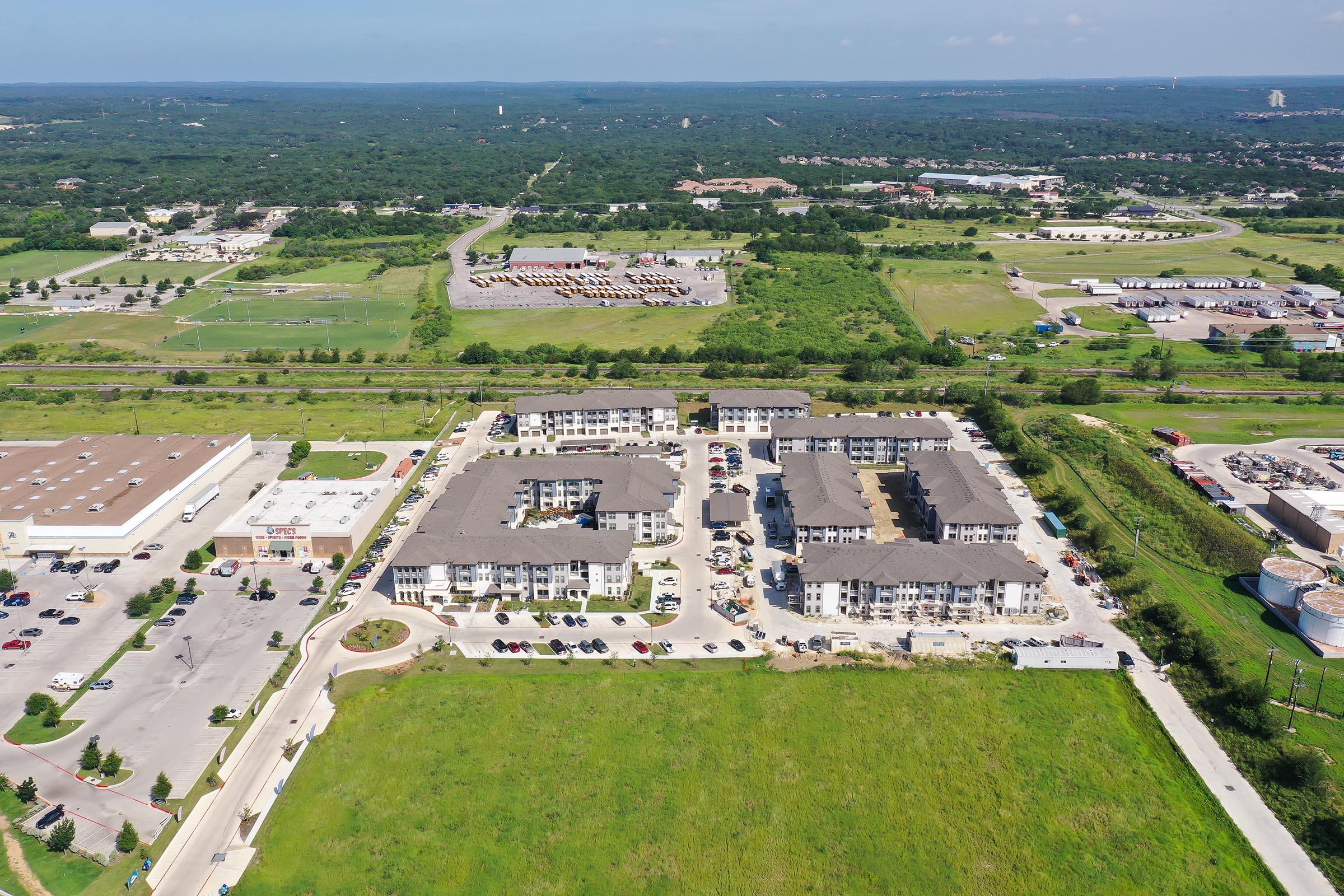 Aerial view of a residential complex surrounded by green fields and commercial areas. The building features a modern design with multiple sections, parking lots, and nearby roads. In the background, additional businesses and open land can be seen, indicating a suburban setting.