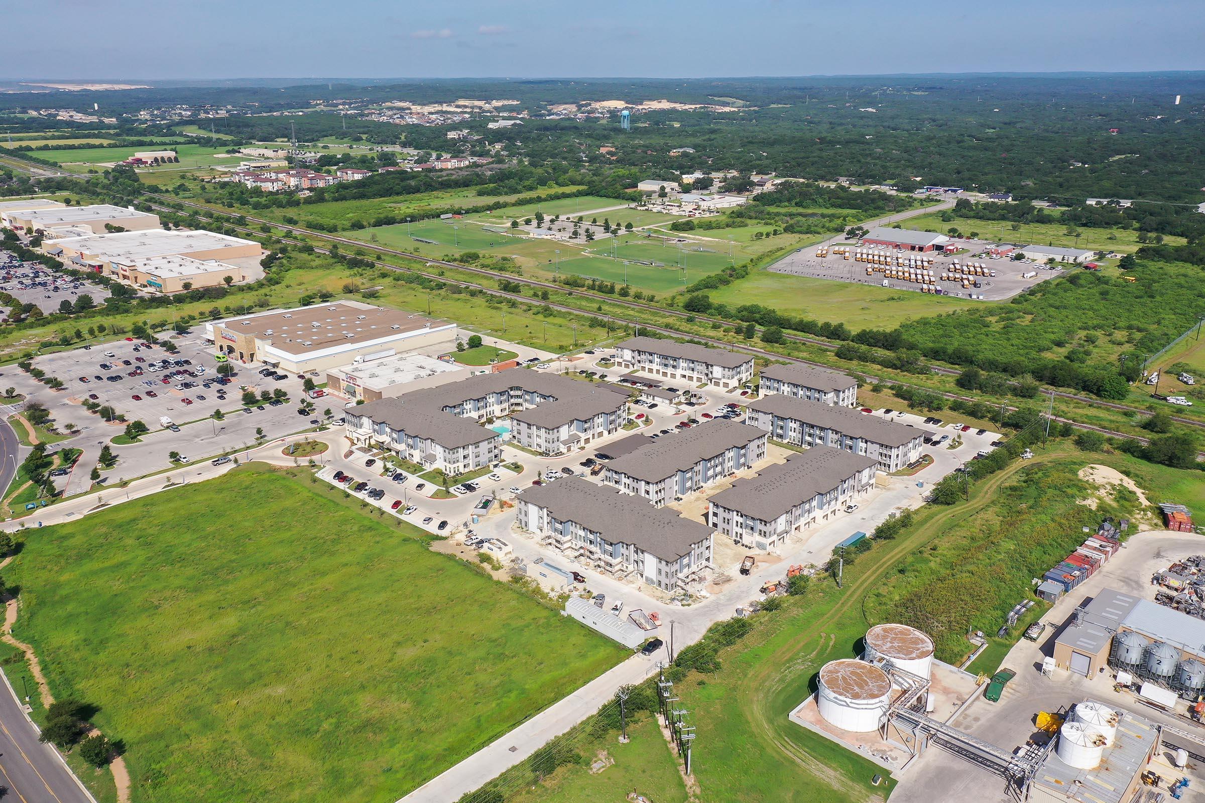 Aerial view of a residential complex surrounded by green spaces and commercial areas. The image shows multiple buildings within the complex, parking lots, and nearby industrial structures, along with roads and fields in the surrounding landscape.