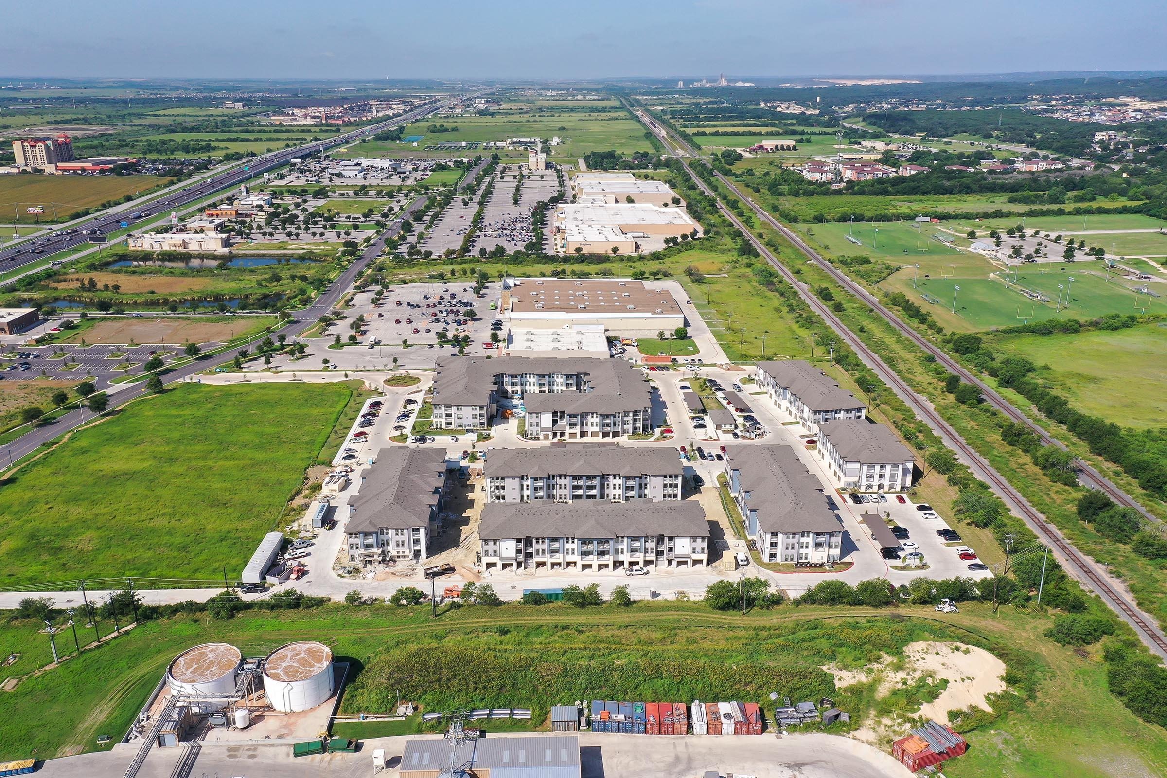 Aerial view of a spacious residential area featuring multiple apartment buildings, surrounded by green fields and parking lots. In the background, a commercial center and a highway are visible, along with a train track and industrial structures. Clear blue skies above.