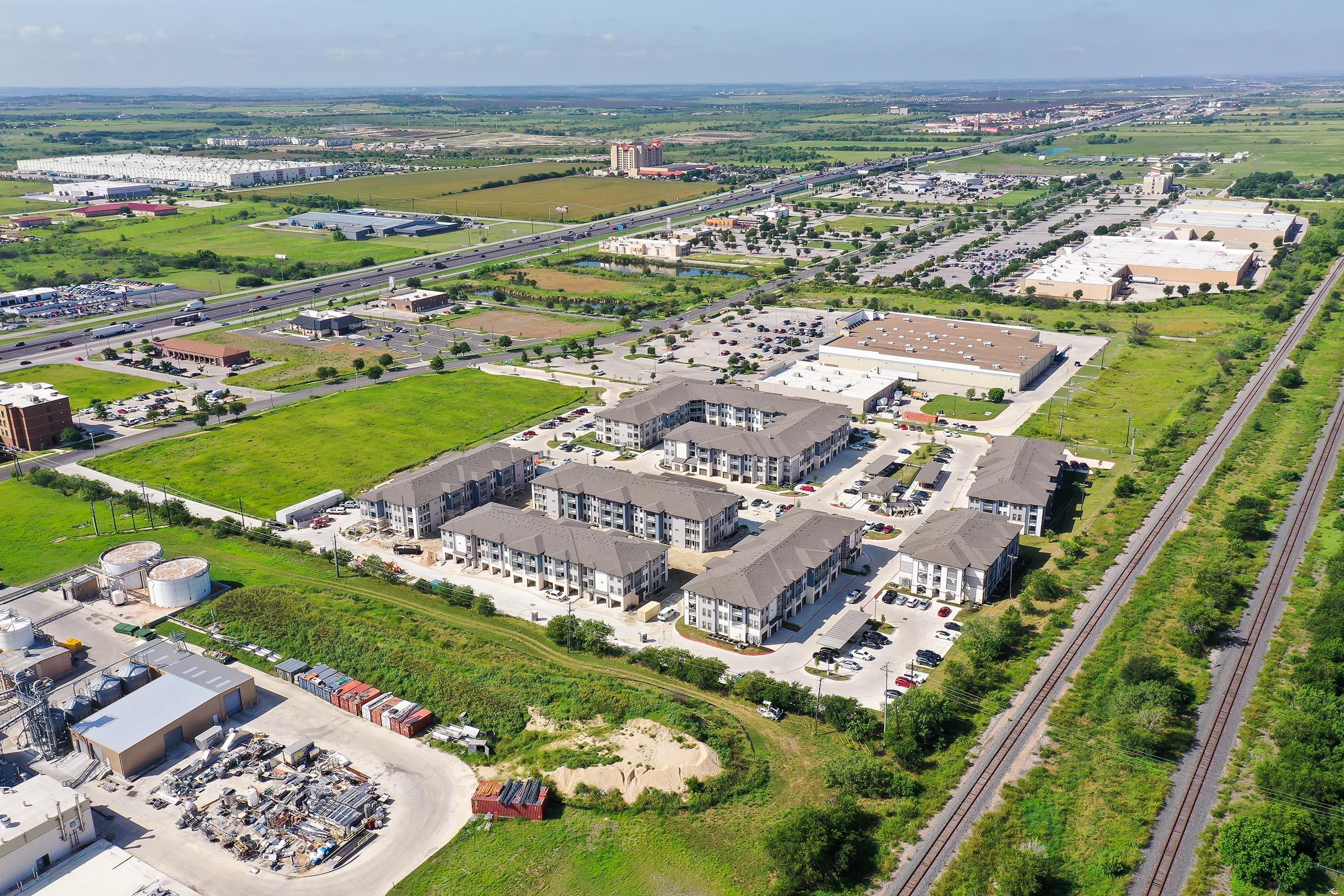 Aerial view of a suburban area featuring new residential apartments, commercial buildings, and parking lots. In the background, highways and expansive green fields are visible, along with a railway line running alongside the buildings. The scene captures a blend of urban development and open land.