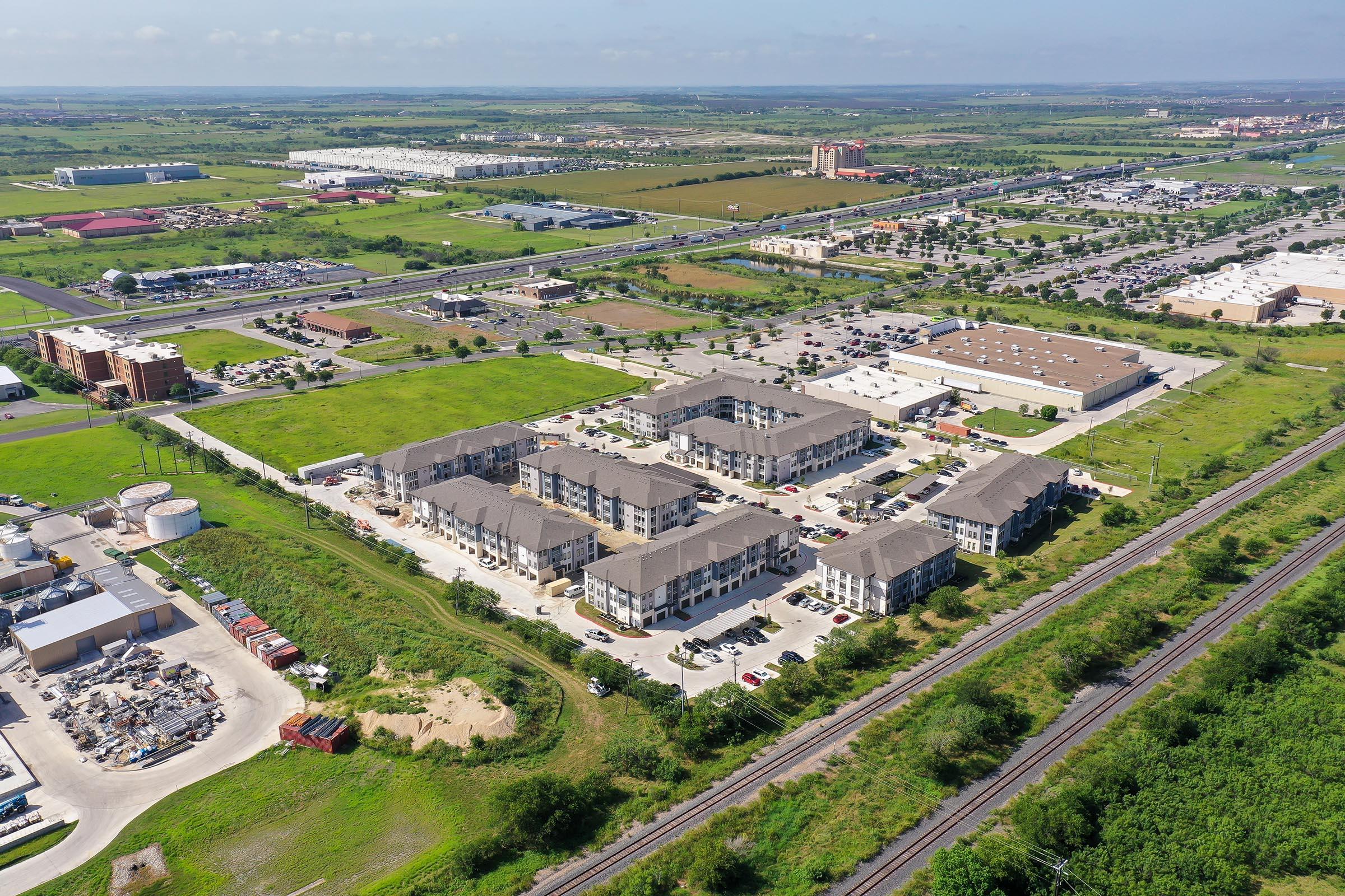 Aerial view of a suburban area featuring a multi-unit residential complex, commercial buildings, and expansive green fields. In the background, there are various parking lots, roads, and a railway line, with a clear blue sky above. The scene shows a blend of developed and natural landscapes.