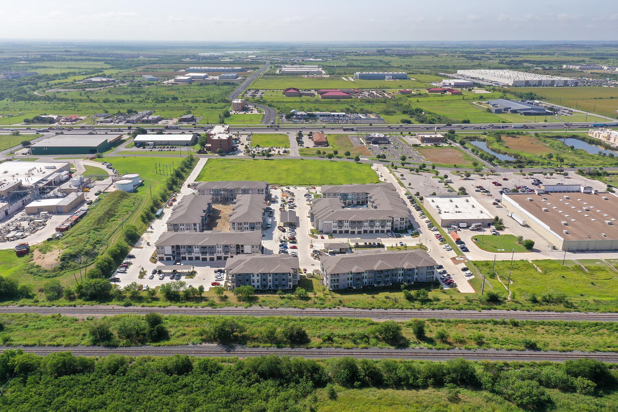 Aerial view of a residential area featuring several apartment buildings surrounded by green spaces, parking lots, and roadways. The background includes commercial buildings and warehouses, along with expanses of open land and infrastructure, indicating a suburban development.