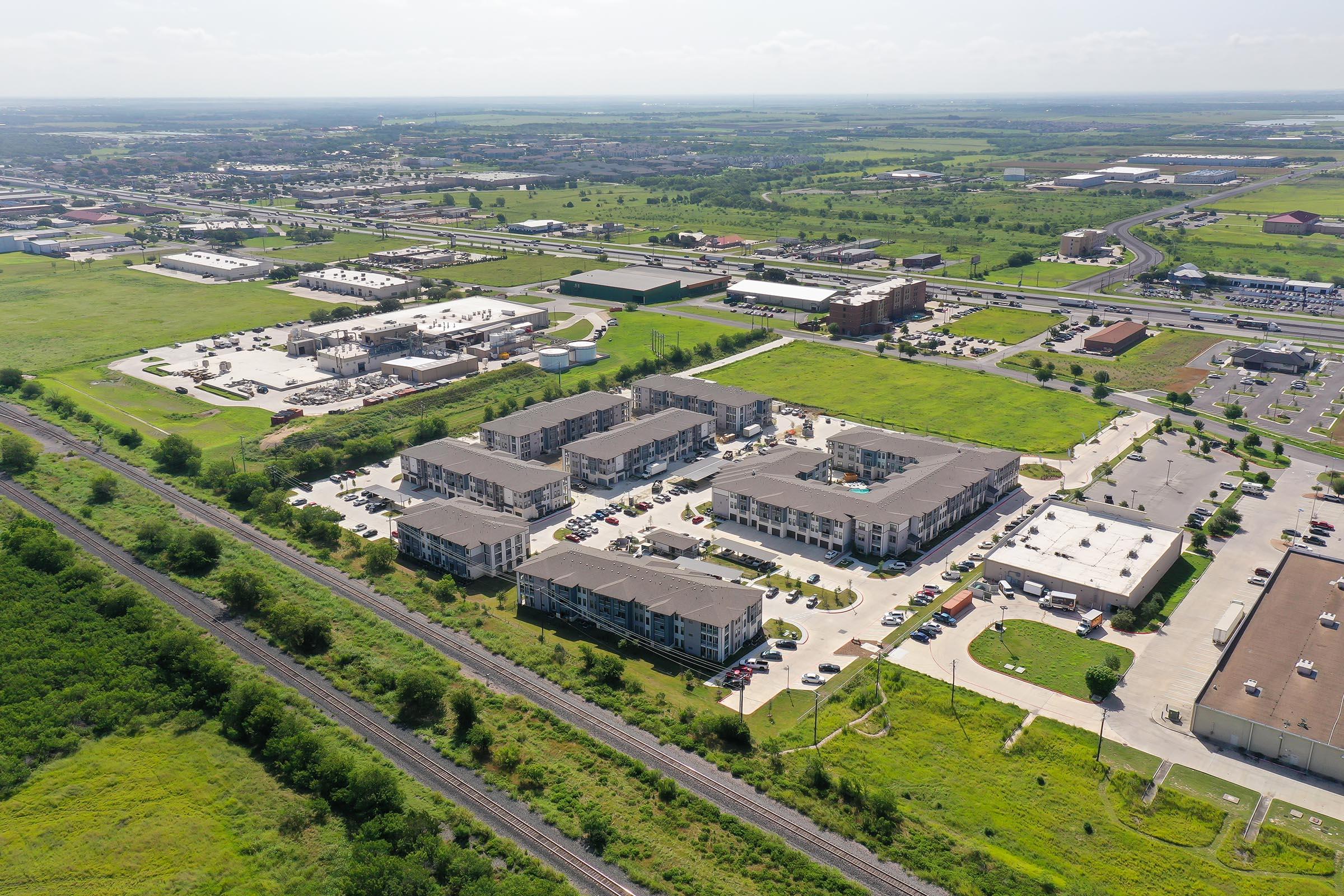 Aerial view of a residential complex surrounded by green fields and commercial buildings. The image showcases several multi-story apartment buildings, a parking area, and adjacent roads, with a vast landscape extending into the distance.