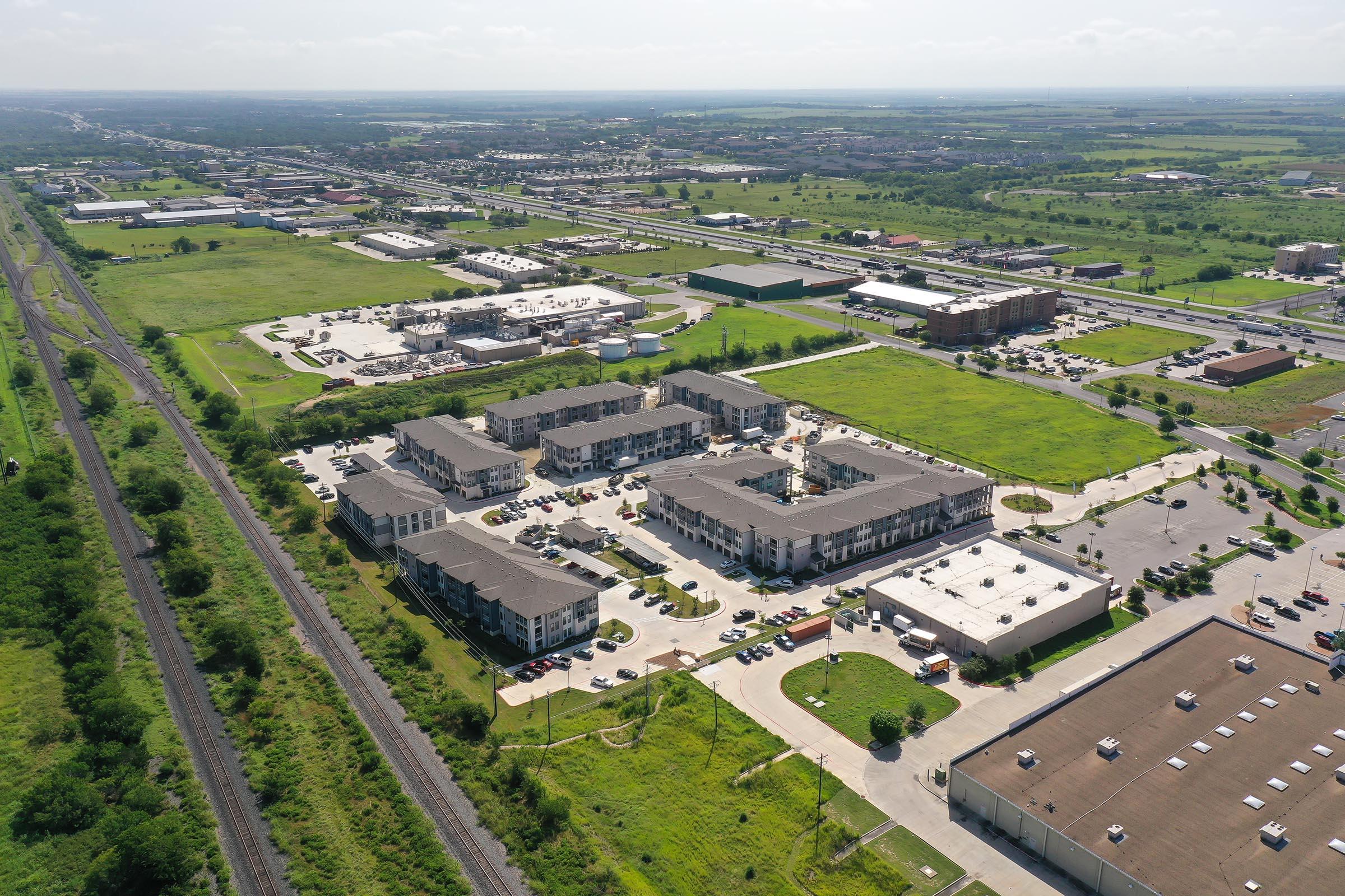 Aerial view of a residential complex surrounded by green fields and commercial buildings. Nearby, there are roads, parking lots, and a railway track. The landscape includes a mix of urban development and open spaces, showcasing a blend of residential and commercial areas.