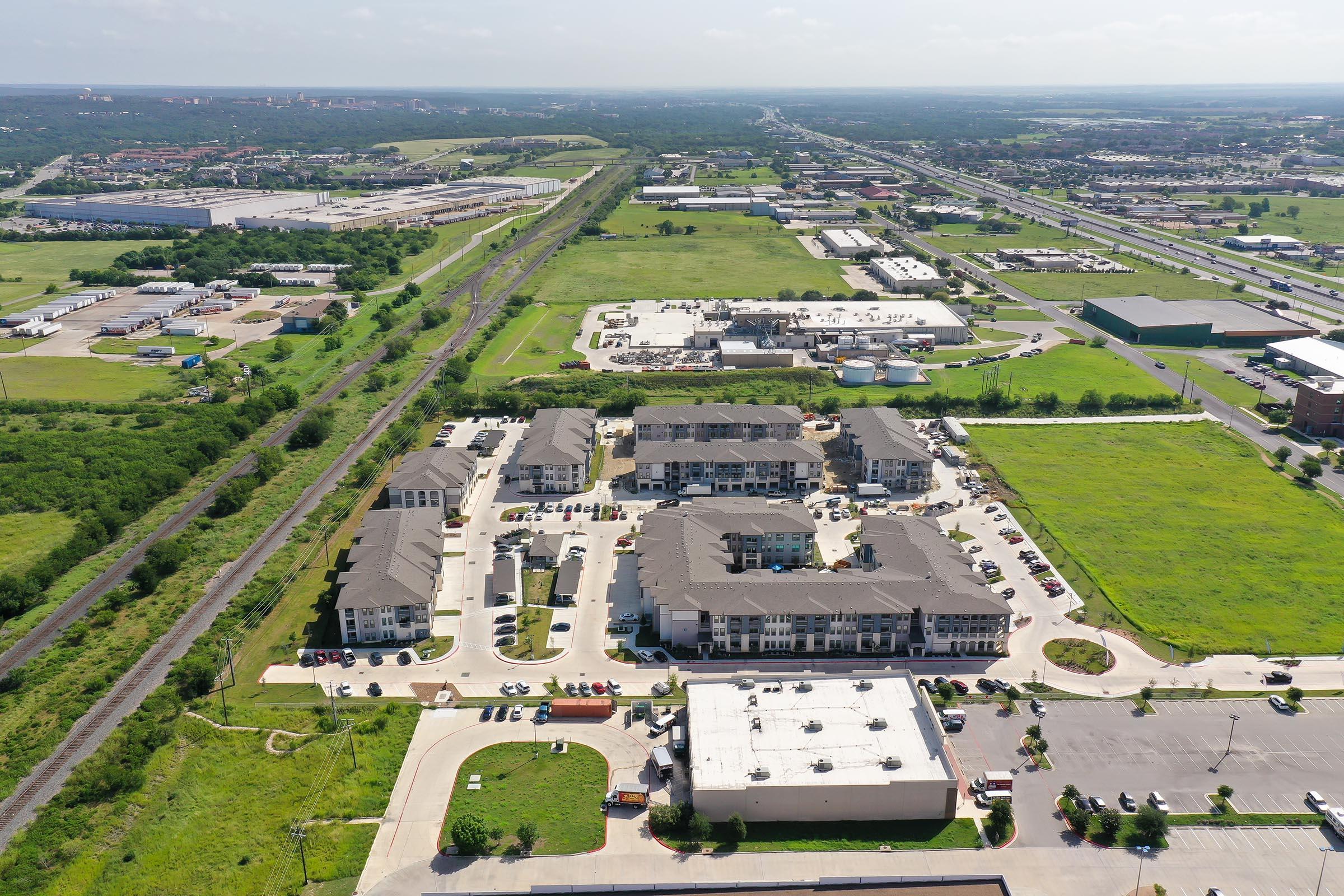 Aerial view of a suburban area featuring a cluster of modern apartment buildings, parking lots, and green open spaces. In the background, there are industrial warehouses and a railway track, with urban development visible in the distance under a clear sky.