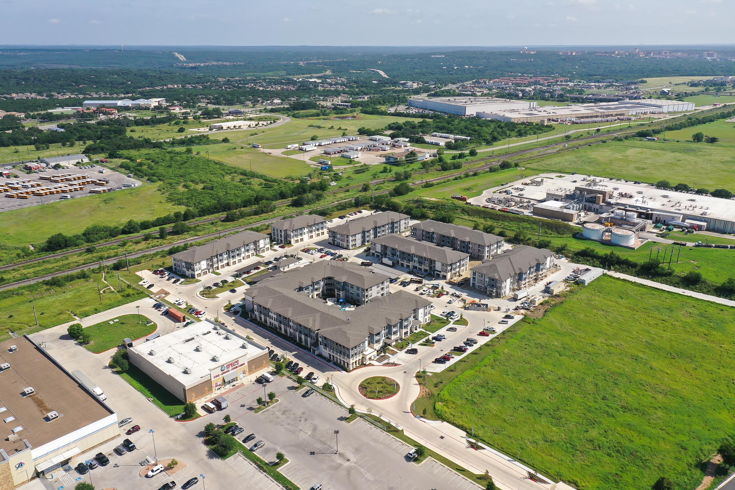 Aerial view of a suburban area featuring a cluster of residential buildings surrounded by green fields and commercial properties. Roads and parking lots are visible, with a distant view of more industrial structures and open land, indicating a blend of urban and rural landscapes.