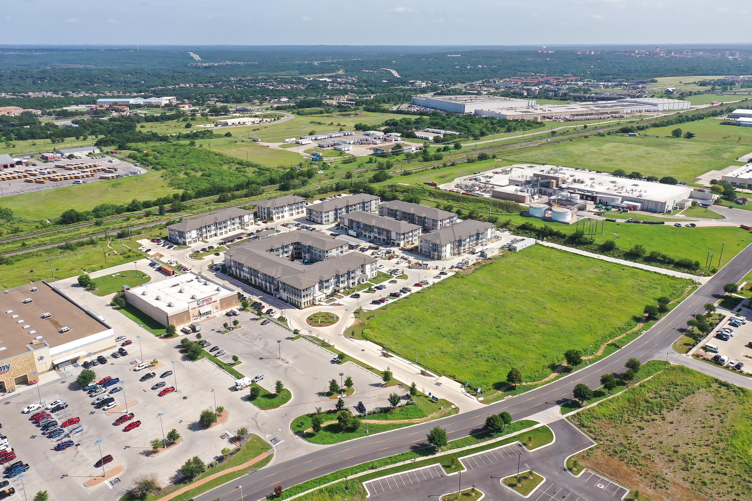Aerial view of a residential area featuring multiple low-rise apartment buildings surrounded by green fields. A parking lot is visible in the foreground, along with nearby commercial buildings and open spaces. The landscape includes patches of trees and distant structures, showcasing a suburban environment.