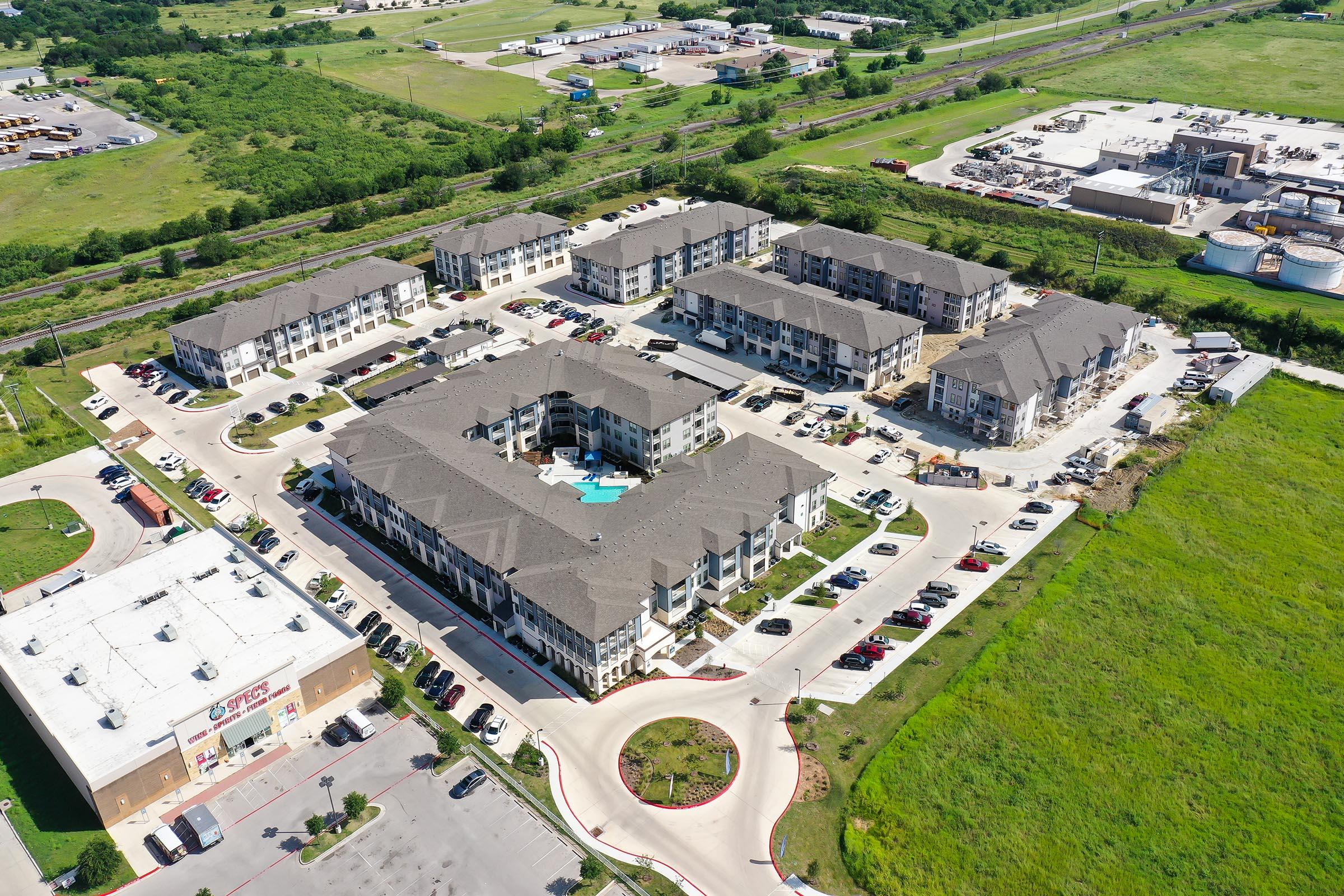 Aerial view of a residential complex featuring multiple apartment buildings with a central pool area, surrounded by parking lots. Nearby commercial buildings and open green spaces can be seen in the background, with highways and roads visible.