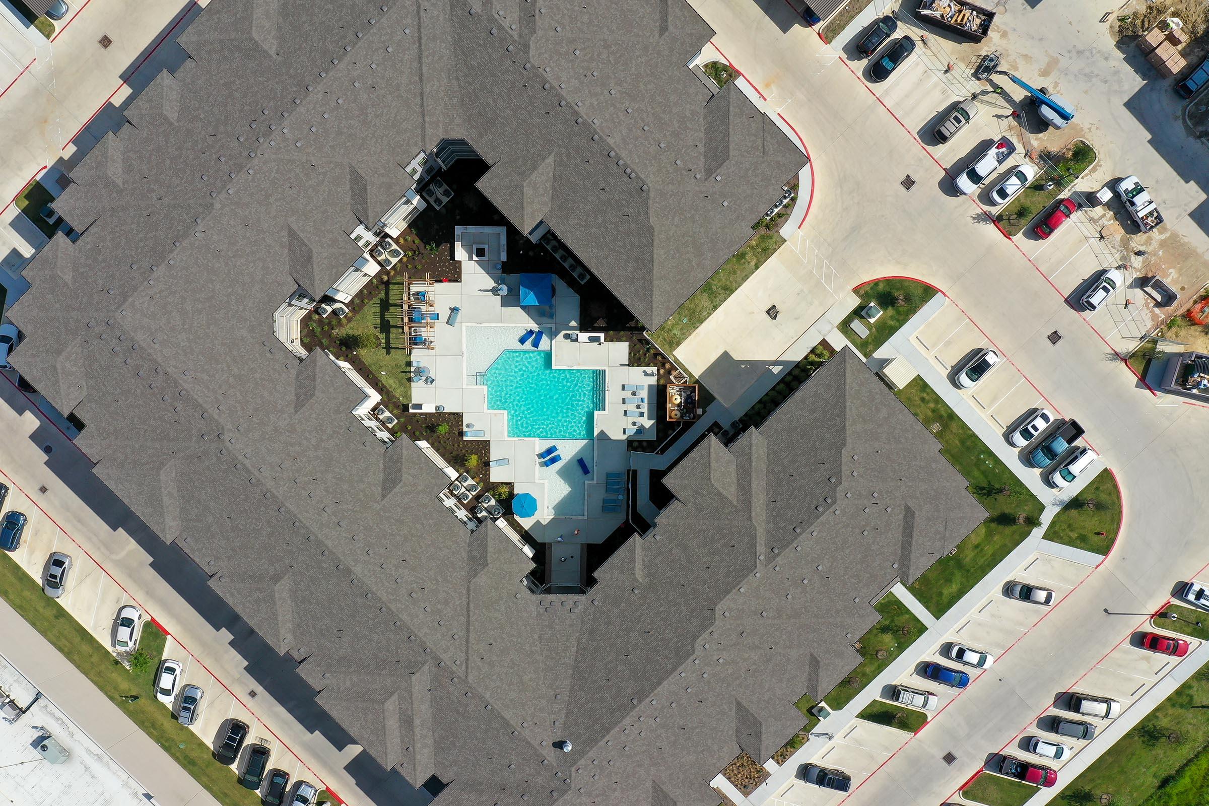 Aerial view of a residential complex featuring a central swimming pool surrounded by lounge areas and landscaping. The complex is surrounded by several parked cars on a circular driveway, with multiple buildings in a symmetrical layout.