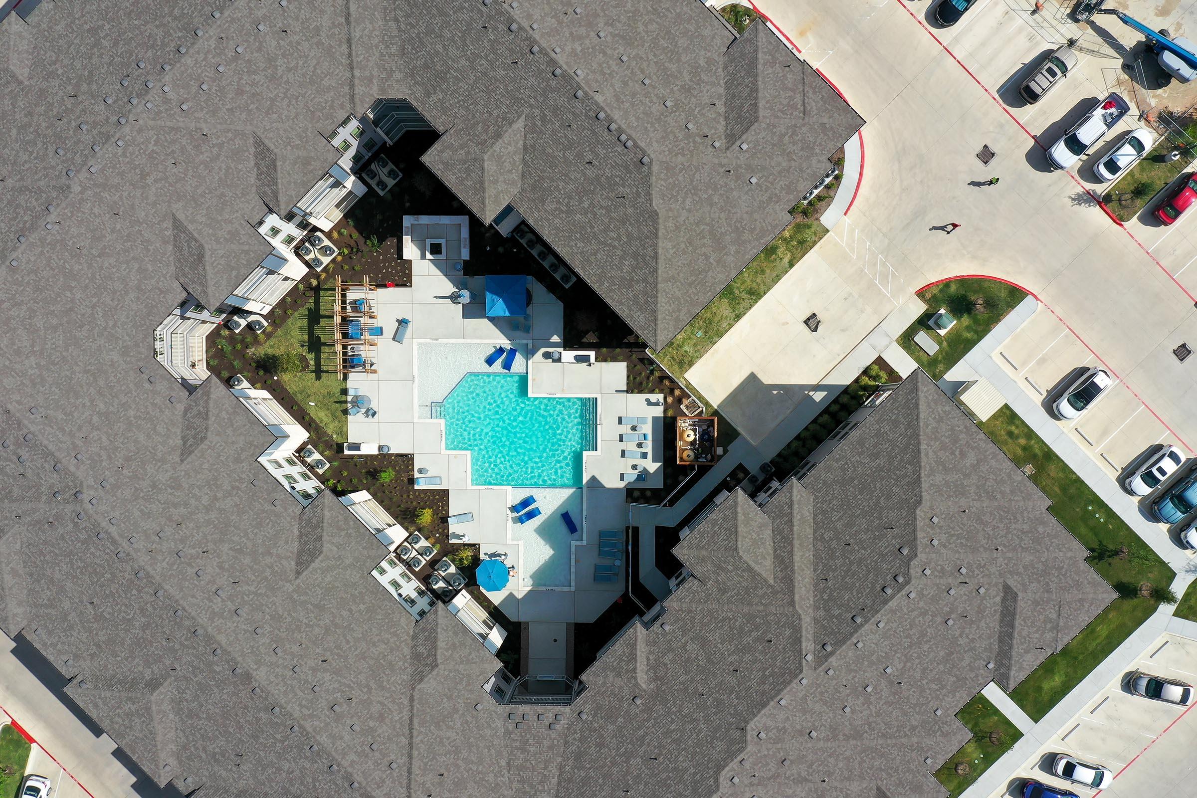 Aerial view of a modern residential complex featuring a central swimming pool surrounded by lounge chairs, landscaped areas, and pathways. The surrounding buildings have dark roofs, and parking lots with cars are visible in the background. The overall layout showcases an inviting outdoor space for residents.