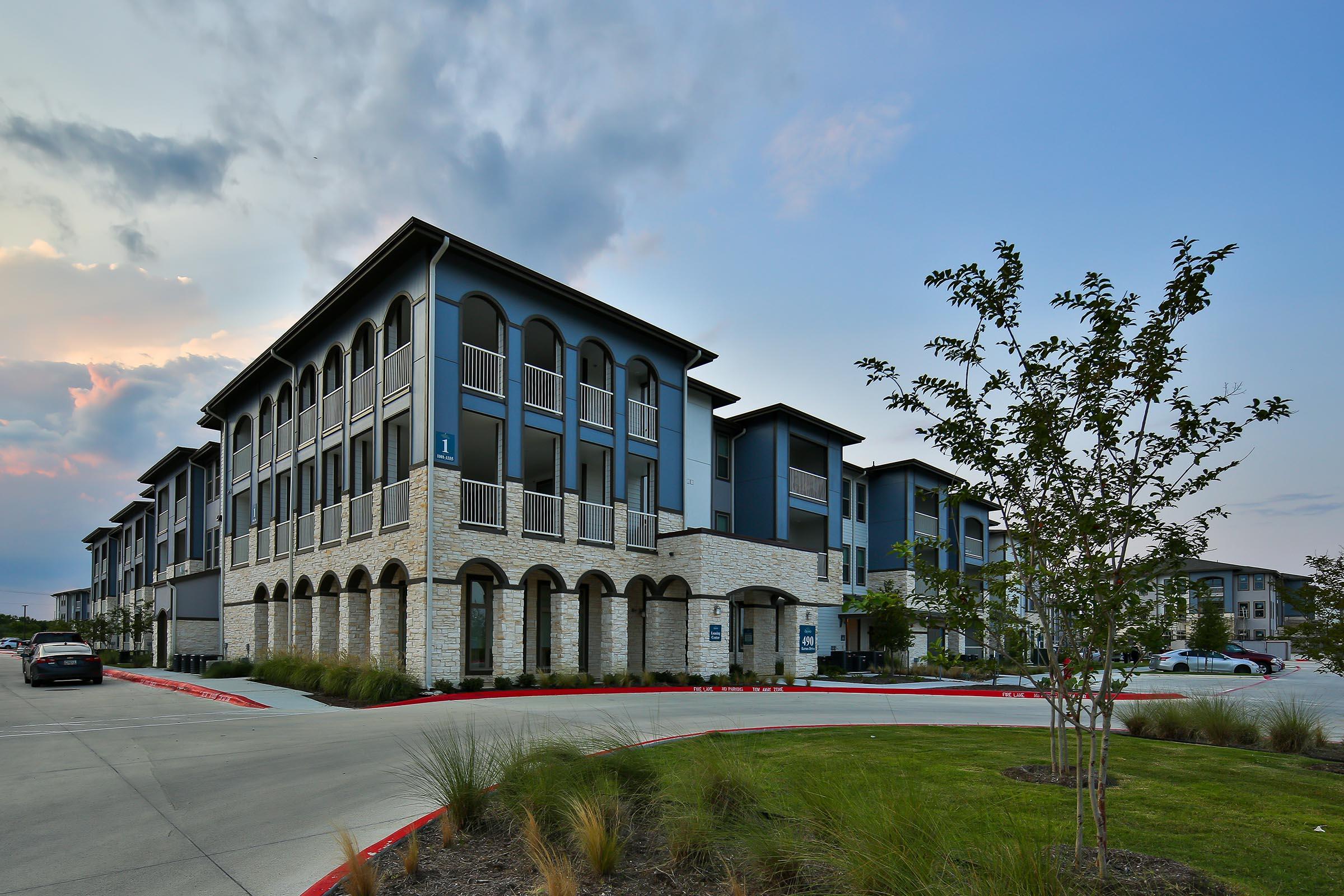 Modern apartment buildings with a mix of stone and blue siding, featuring balconies and arched entrances. The setting includes landscaped greenery and a circular drive, with a partly cloudy sky in the background.