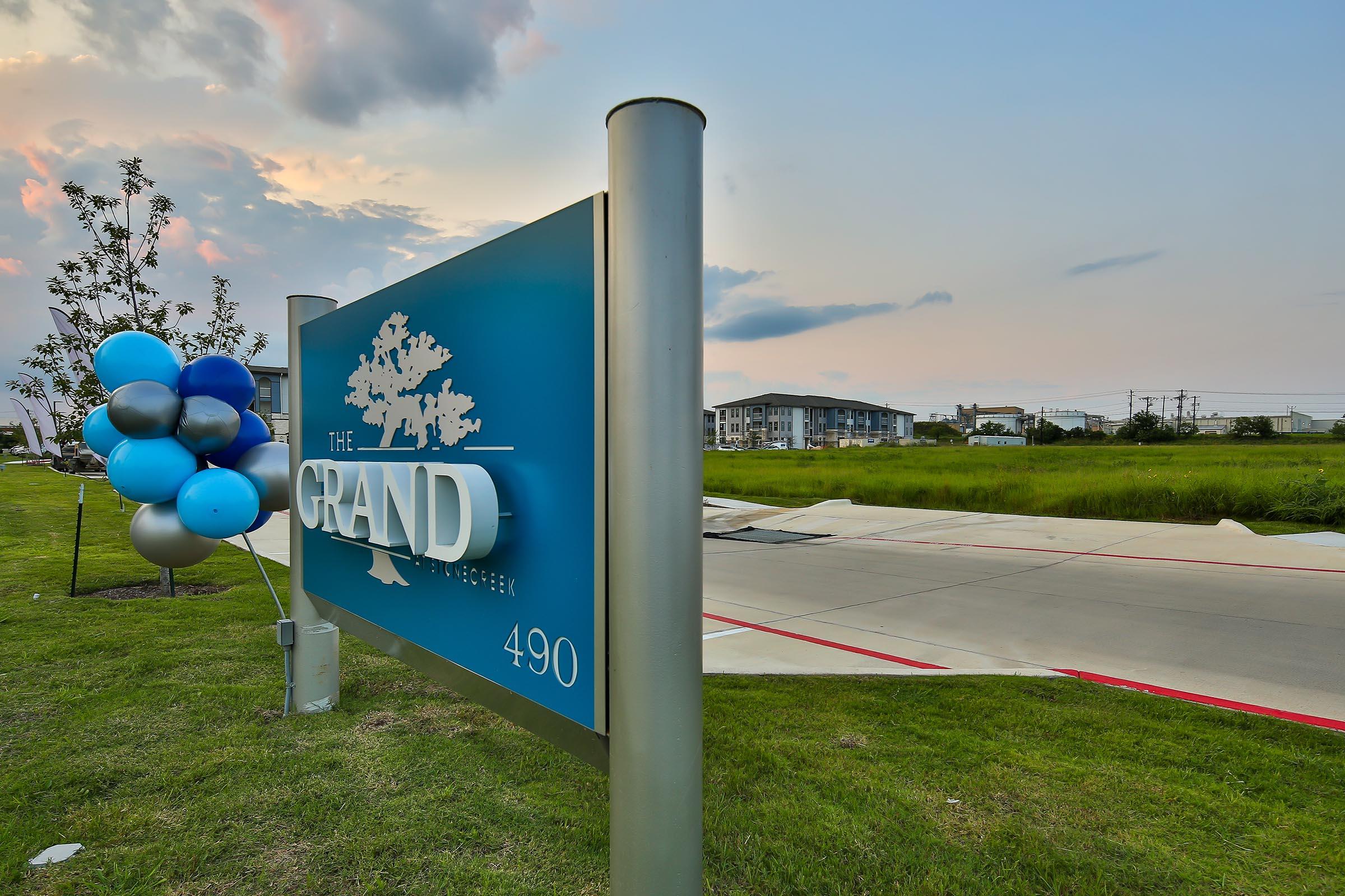 Sign for "The Grand" at 490, surrounded by a grassy area with balloons in shades of blue and silver. The sky features soft clouds at sunset, creating a serene atmosphere. In the background, buildings can be seen along with power lines.