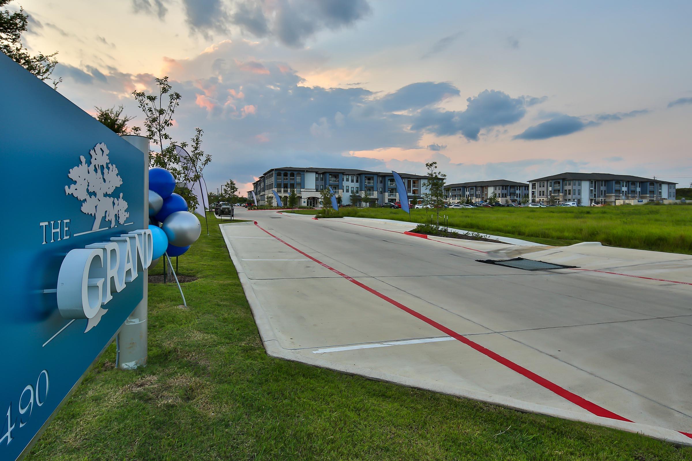 Sign for "The Grand" with blue balloons in foreground, leading to an entrance road. In the background, modern apartment buildings under a cloudy sky during sunset. Green grass along the roadside enhances the inviting atmosphere of the property.