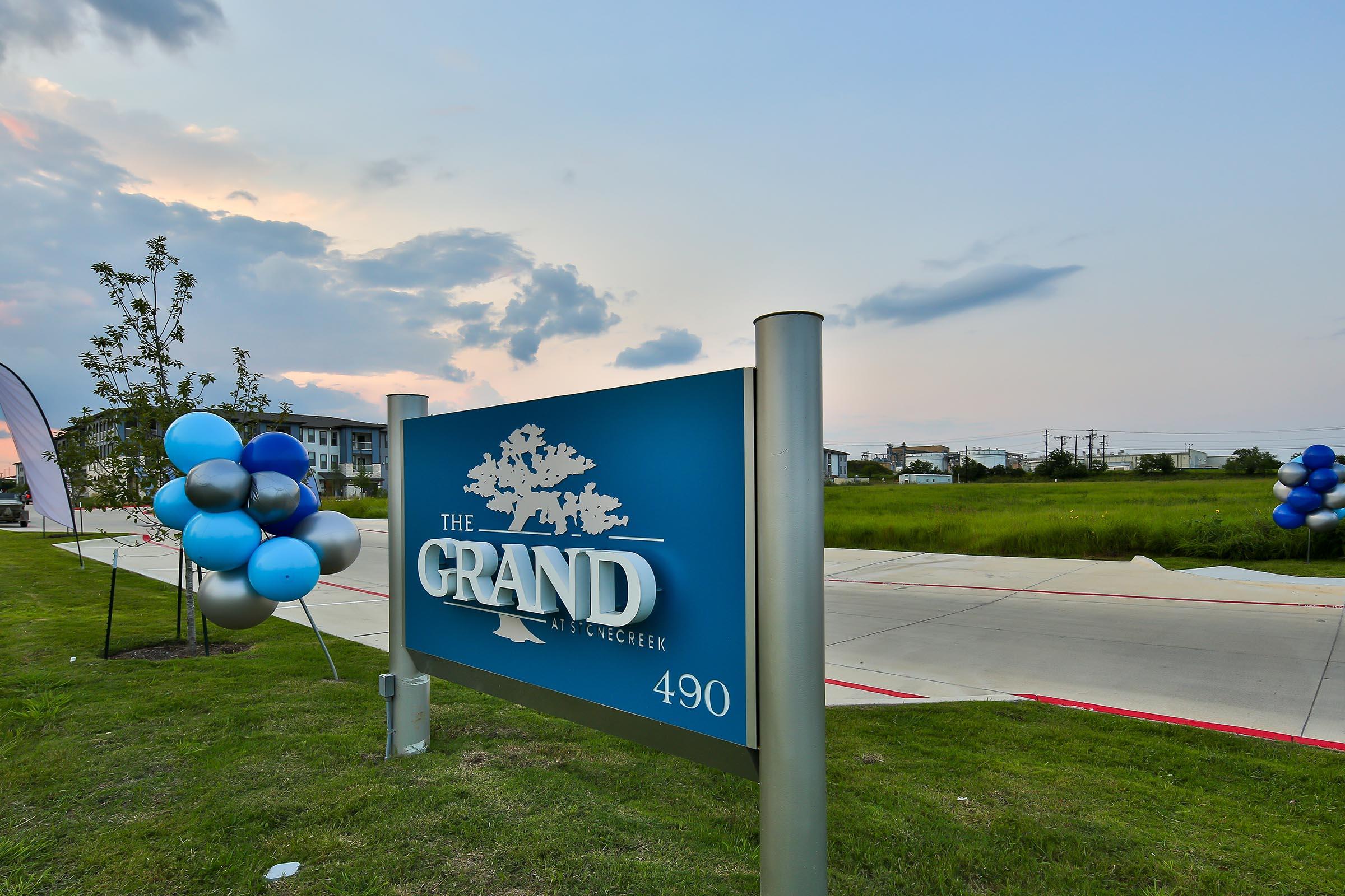 Sign for "The Grand" located at 490, with a blue and white color scheme. In the foreground, there are decorative balloons in shades of blue and silver. The background features a grassy area with a cloudy sky, indicating late afternoon or early evening.
