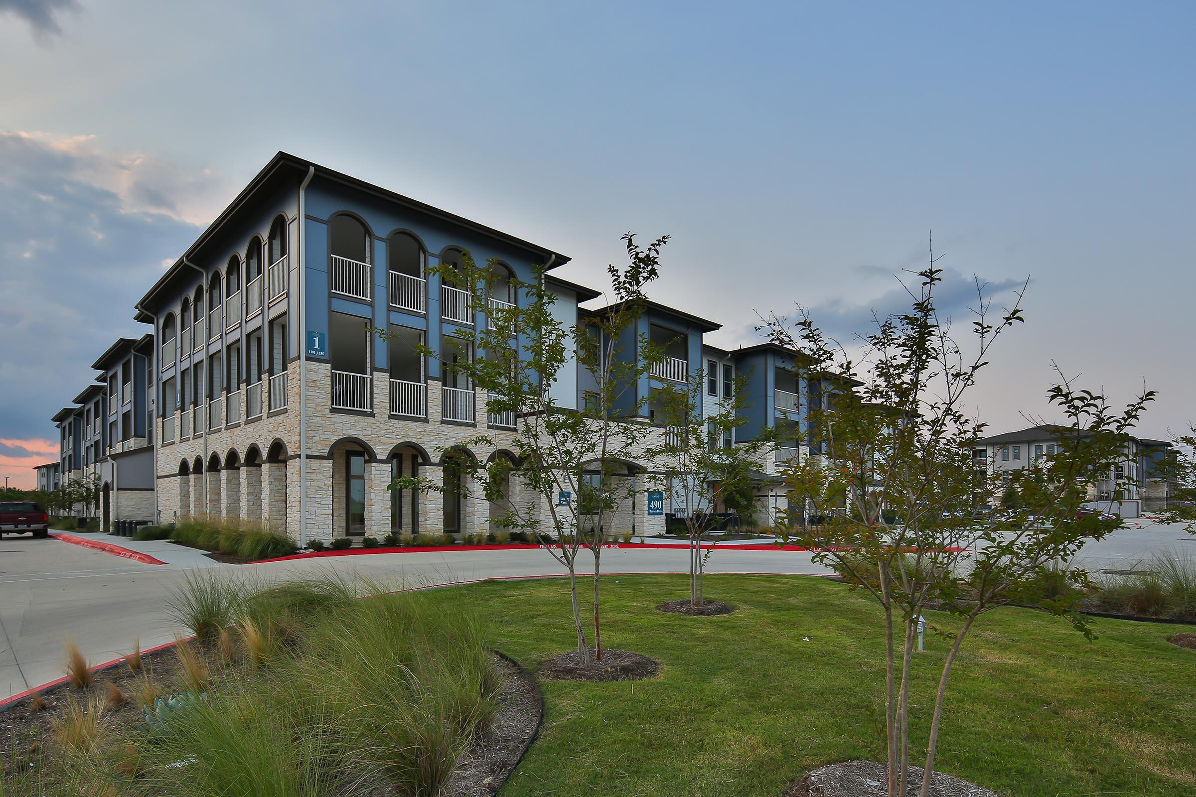 A modern multi-story building with a stone façade, featuring balconies and large windows. The structure is surrounded by landscaped grass and small trees. The scene is set during dusk, with a colorful sky in the background. There is a paved driveway leading into the entrance of the building.