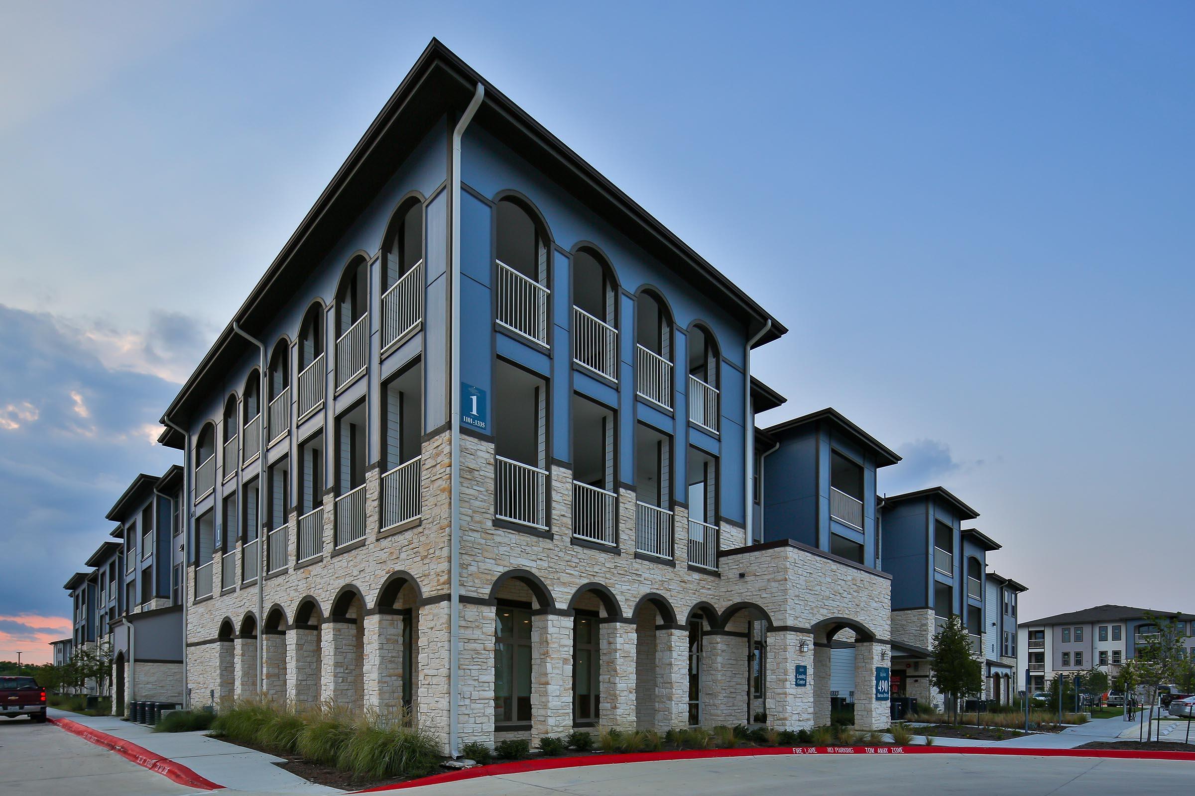 Modern multi-story residential building with a combination of stone and gray exterior. Features large windows, arched entrances, and a landscaped area. The setting is a contemporary neighborhood, with several buildings visible in the background under a blue sky.