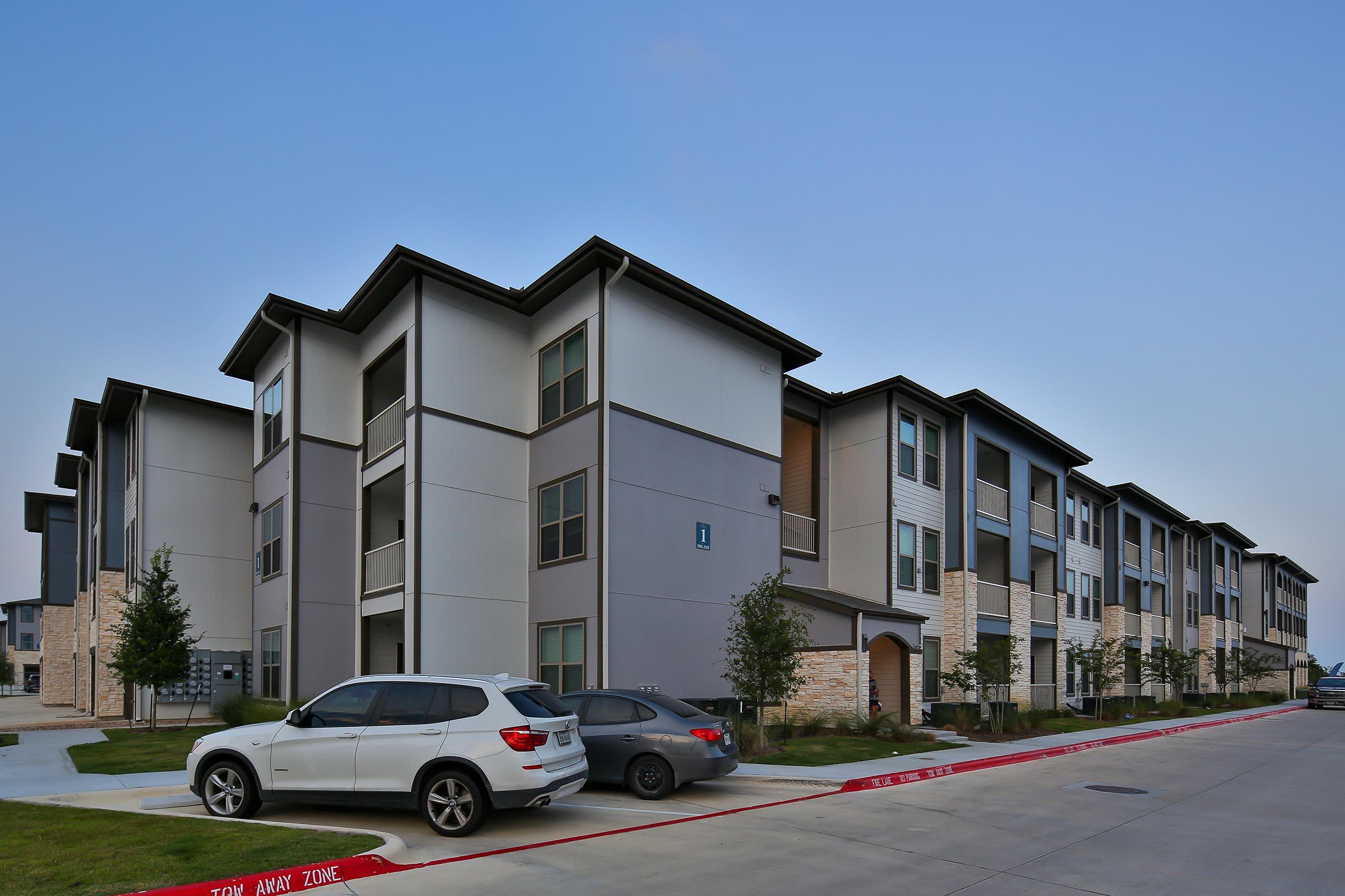 A modern apartment complex with three-story buildings featuring a mix of gray and beige siding. The foreground shows a parking area with two vehicles parked beside landscaped areas. The sky is clear and blue, and the setting appears to be a residential neighborhood.