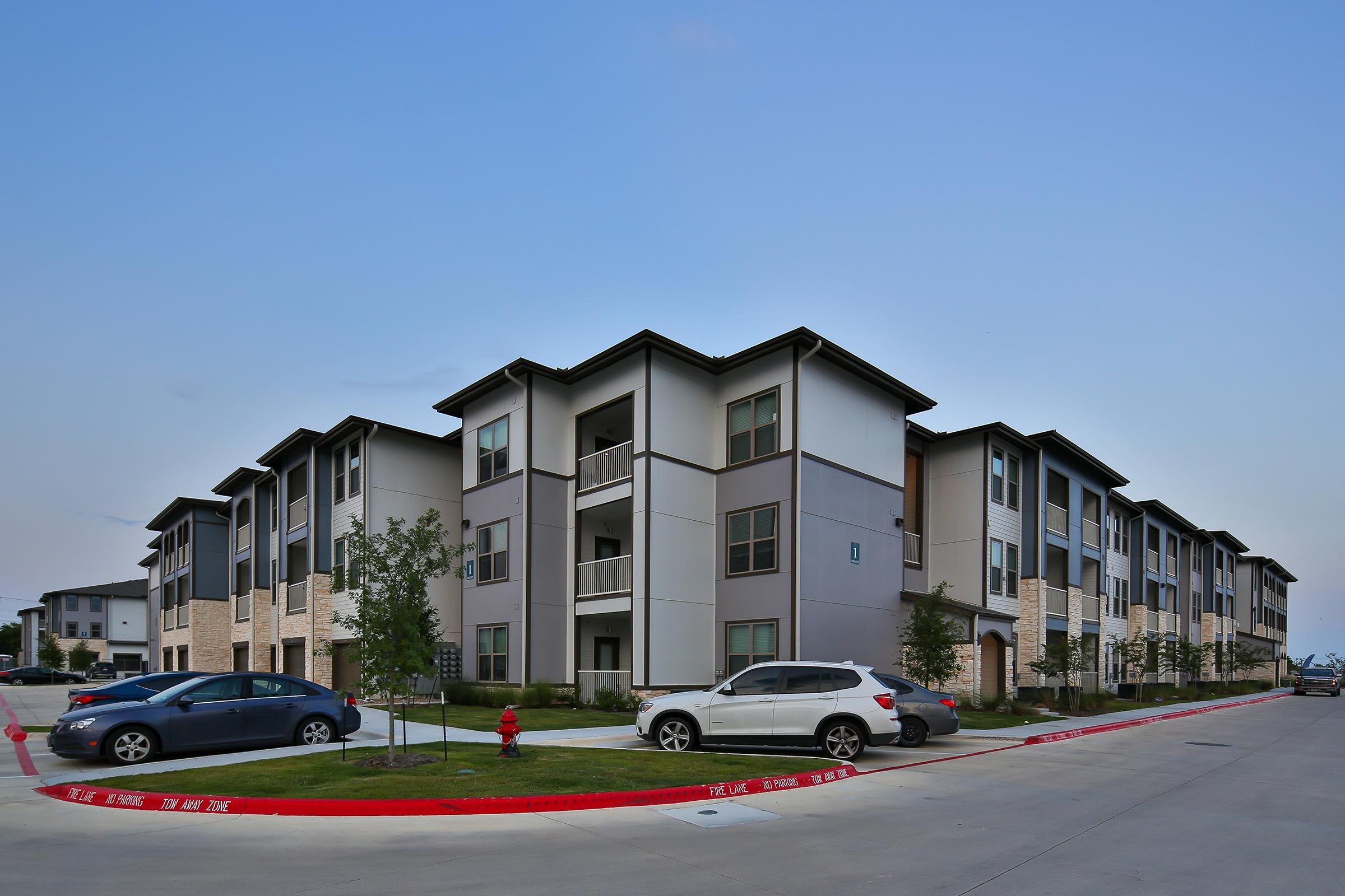 A modern apartment complex featuring multiple three-story buildings with a mix of stone and siding exteriors. The landscape includes well-maintained grass and shrubs, with parked cars and a fire hydrant visible along the driveway. The sky is clear and blue, indicating a sunny day.