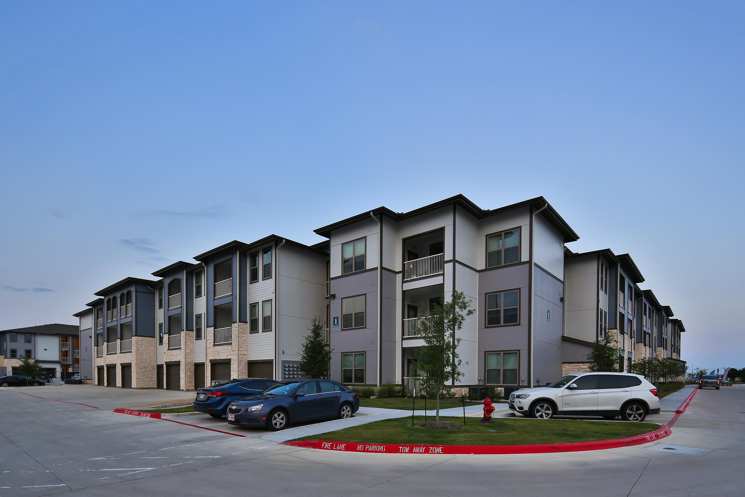 Modern apartment complex with a mix of two and three-story buildings. The buildings have a contemporary design with balconies and large windows. A parking lot in front features several parked cars. The surrounding area includes well-maintained green spaces and trees under a clear blue sky.