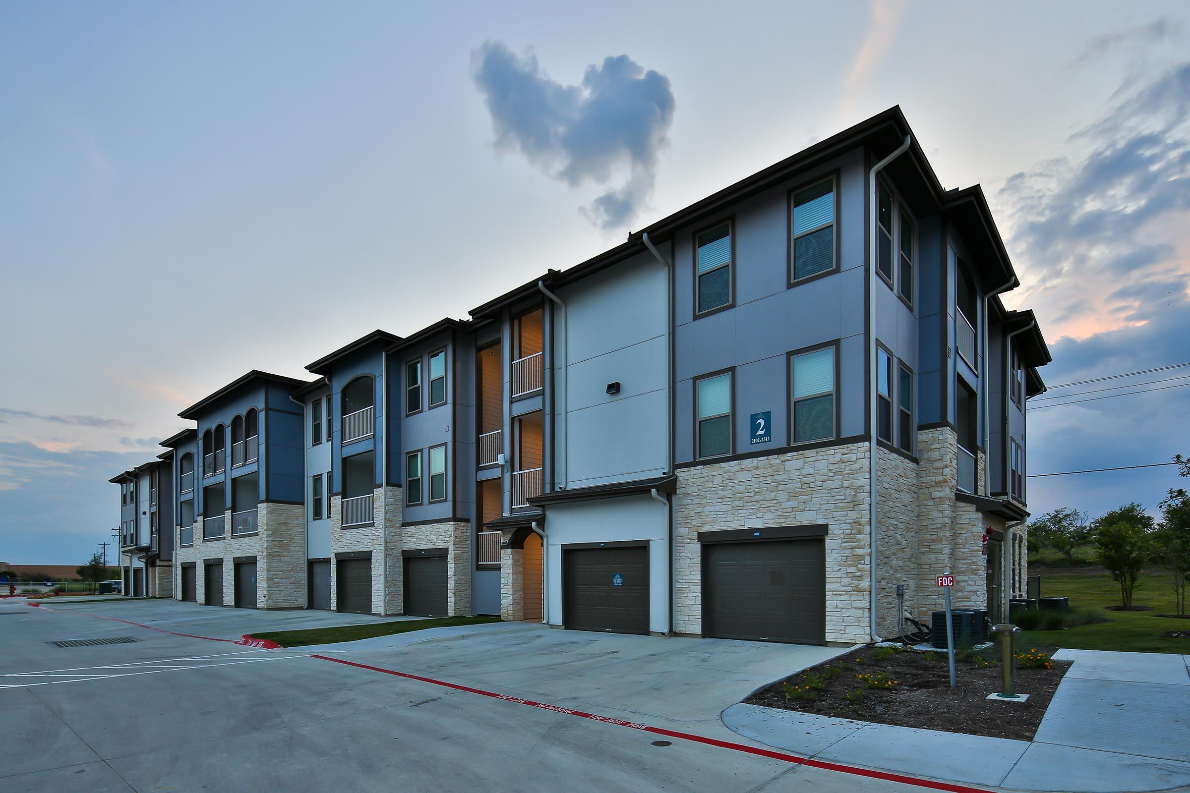Modern multi-unit residential building featuring a combination of grey siding and natural stone elements. The structure has multiple floors, with a parking area below. The sky is partly cloudy, hinting at dusk.