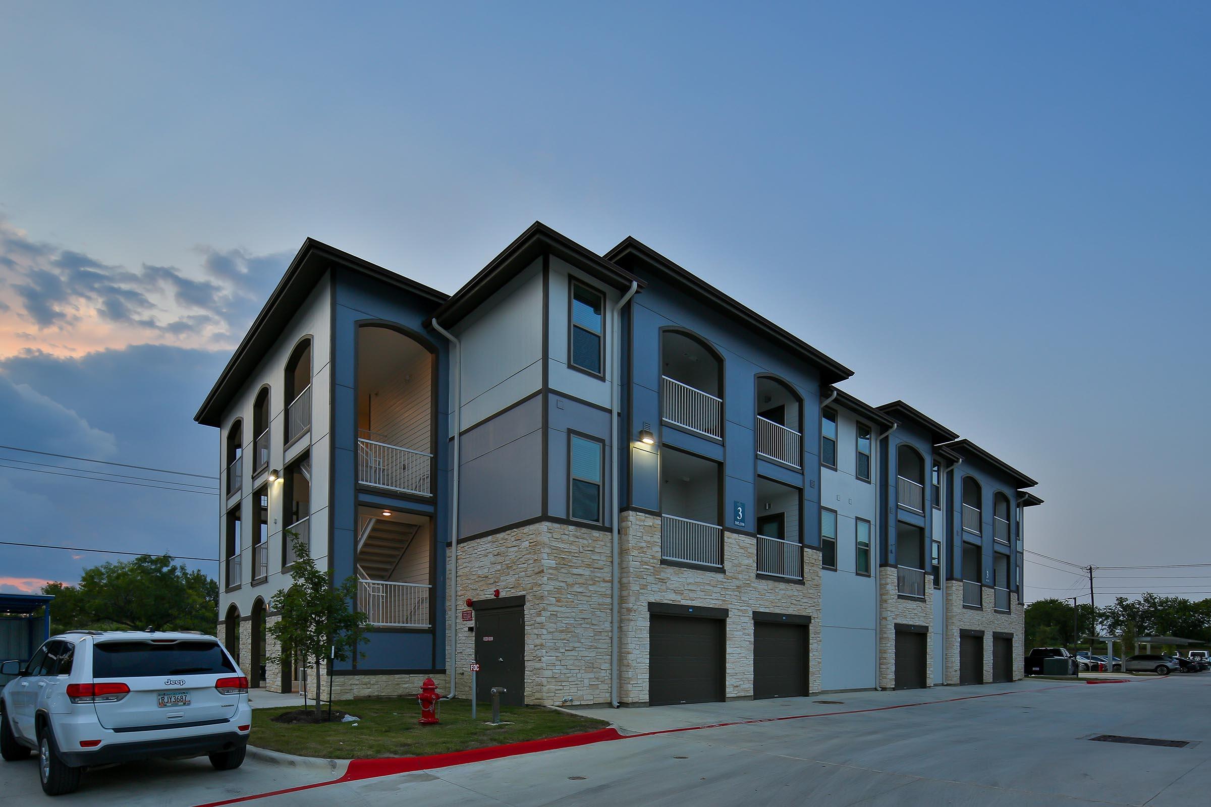 A modern multi-unit apartment building at dusk, featuring a light-colored façade with dark accents, balconies, and multiple garage doors. The scene is serene, with a light cloud cover and trees nearby, highlighting the contemporary design of the architecture.