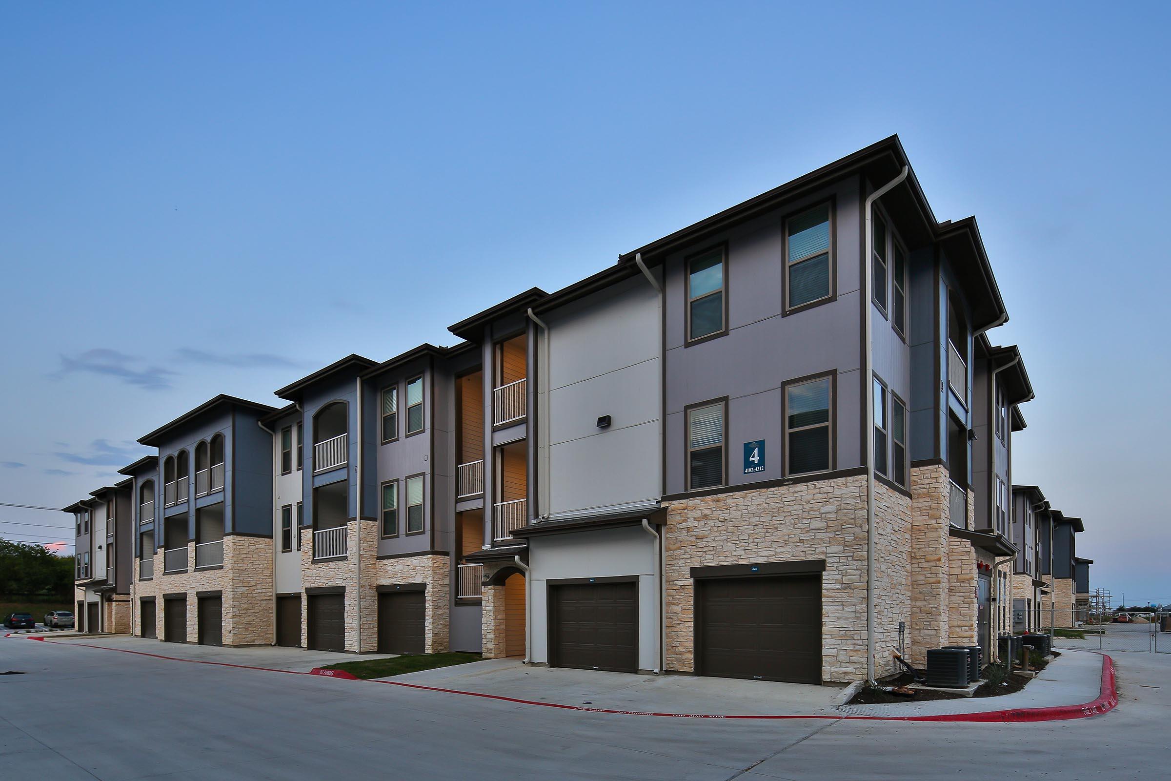 Modern multi-unit residential buildings with a mix of stone and siding exteriors. The three-story structures feature garages on the ground floor and balconies on the upper levels, set against a clear evening sky. The layout includes a paved parking area and landscaping elements.