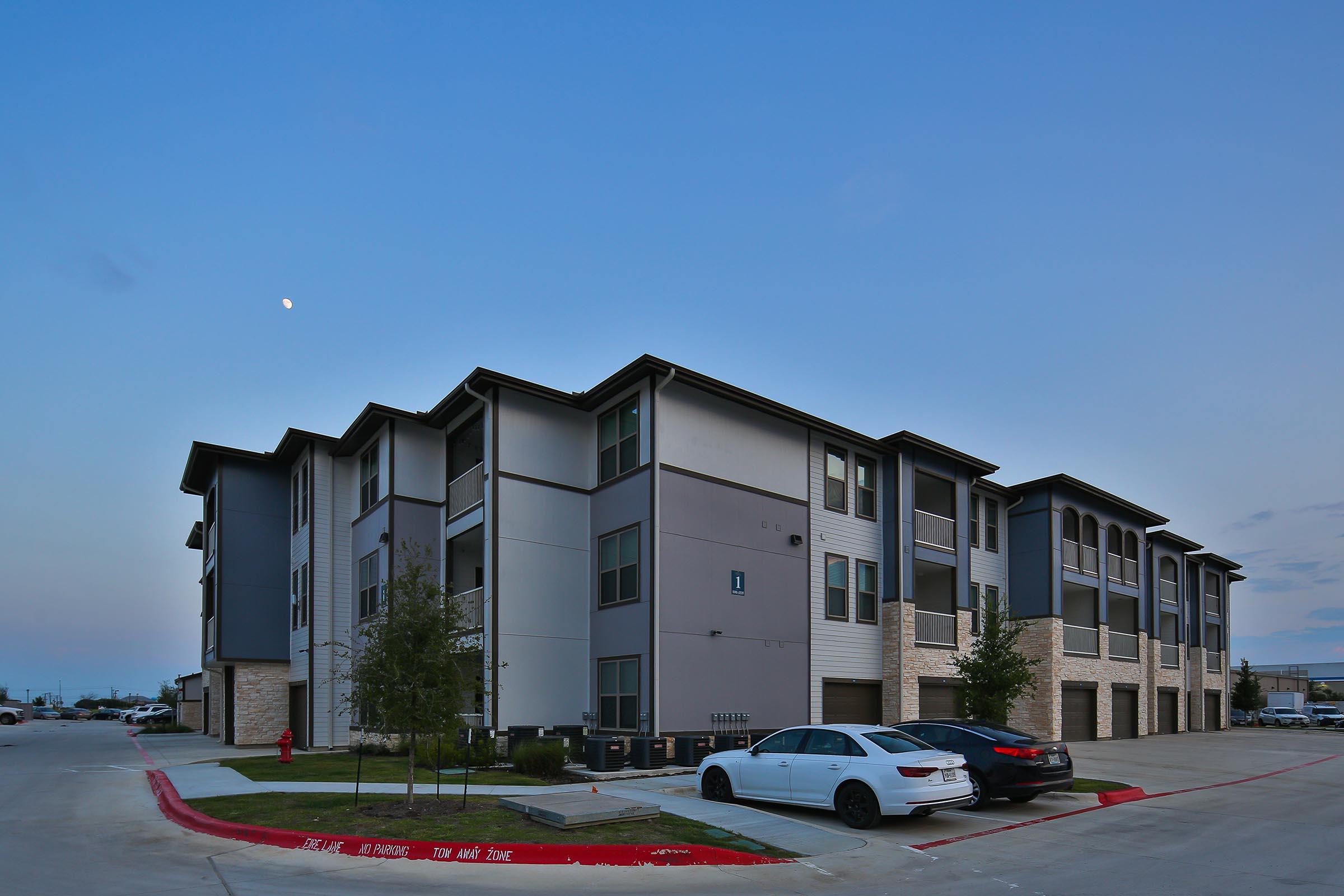 A modern three-story apartment building with a mix of dark and light siding, surrounded by a paved parking area. Several vehicles are parked in front, and small trees are planted along the sidewalk. The sky is transitioning into evening with a visible moon.