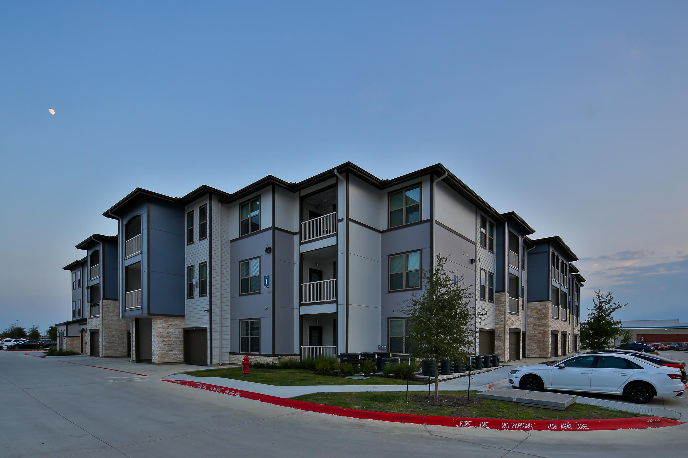 Modern multi-story apartment building with a mix of gray and cream exterior. There are balconies, large windows, and landscaping featuring small trees and grass. A white car is parked in the foreground, and the sky is transitioning to dusk with a visible moon.