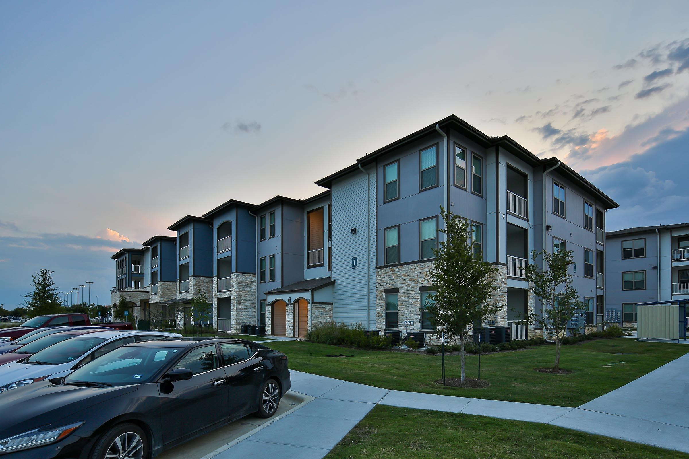 A modern residential building with multiple stories, featuring a mix of gray and beige siding. In the foreground, there are several parked cars along a well-maintained grassy area, with trees and a pathway leading to the entrance. The sky is partly cloudy, hinting at dusk.