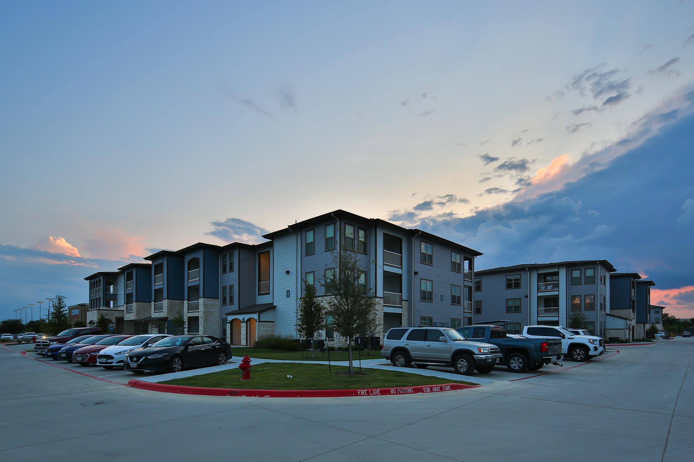 Modern apartment complex with multiple units, parked cars in the foreground, and a colorful sunset sky in the background. The buildings feature a contemporary design with a mix of grey and blue colors, surrounded by green landscaping and a clear pathway.