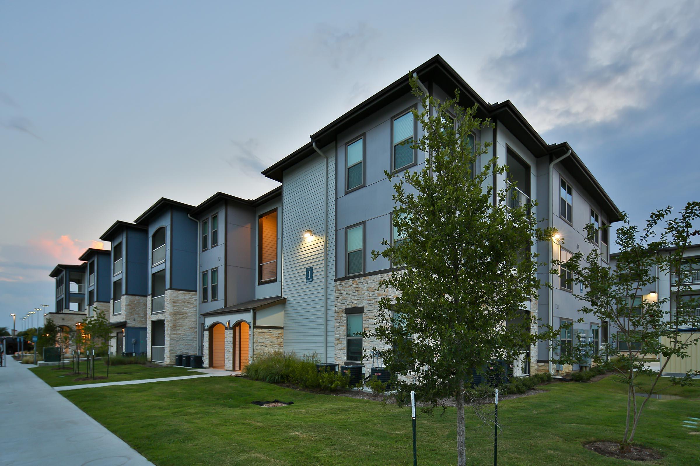 A modern multi-story apartment building with a landscaped yard, featuring minimalistic architecture and large windows. The exterior combines gray and beige tones with stone accents. Soft lighting highlights the building, and the sky shows hints of sunset with scattered clouds.