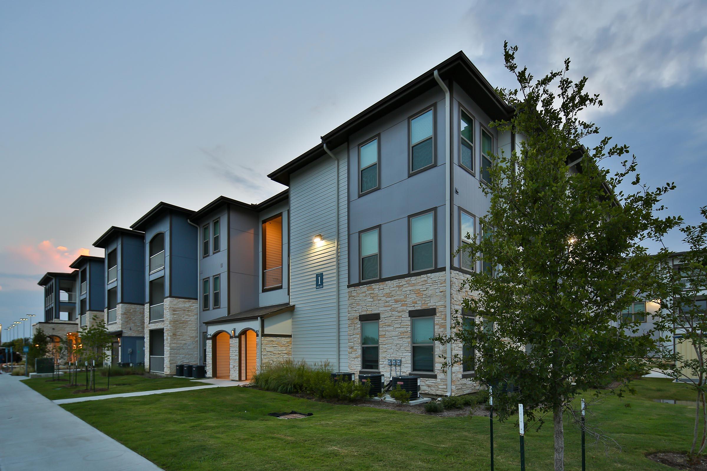 Three-story modern apartment building with a mix of gray and beige siding, featuring large windows. The exterior is lined with small trees and landscaping. The setting is during twilight with soft lighting from the building's windows, and a sidewalk runs along the front.