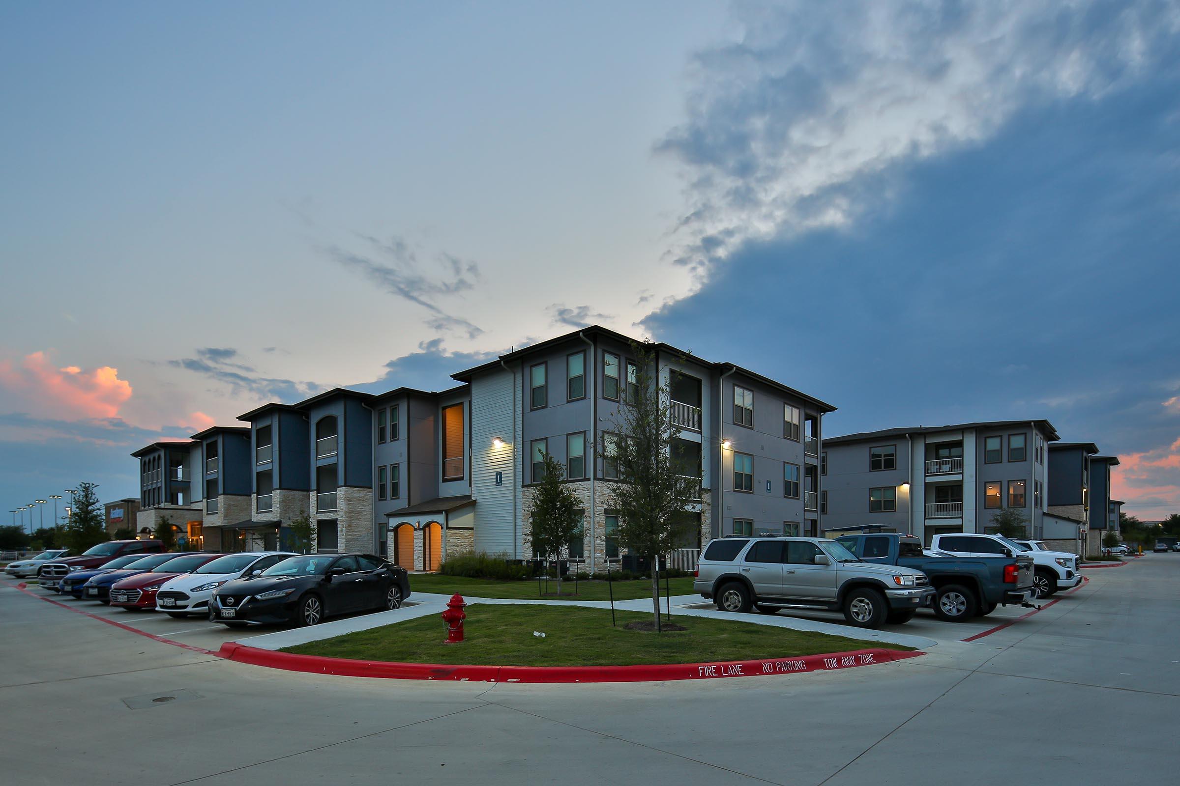 Modern residential apartment complex at dusk, featuring multiple two- and three-story buildings with a mix of colors. A parking lot is filled with various vehicles, and landscaping includes small trees and grass. The sky is partly cloudy, adding a serene atmosphere to the scene.