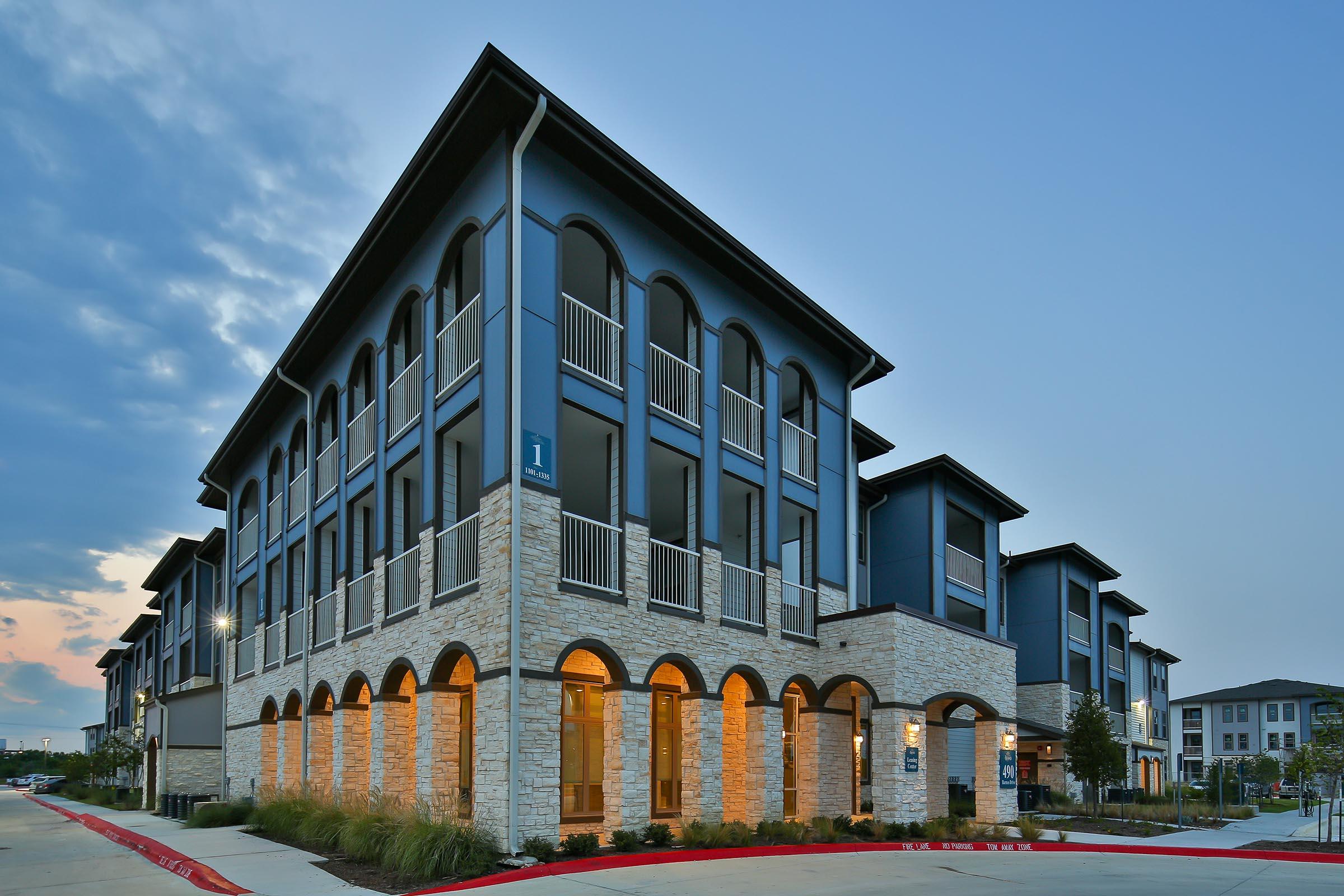 Modern multi-story apartment building featuring a mix of stone and blue-grey exterior. The design includes arches on the ground level, with warm lighting at dusk. Lush landscaping surrounds the property, and the sky is softly lit, indicating early evening.