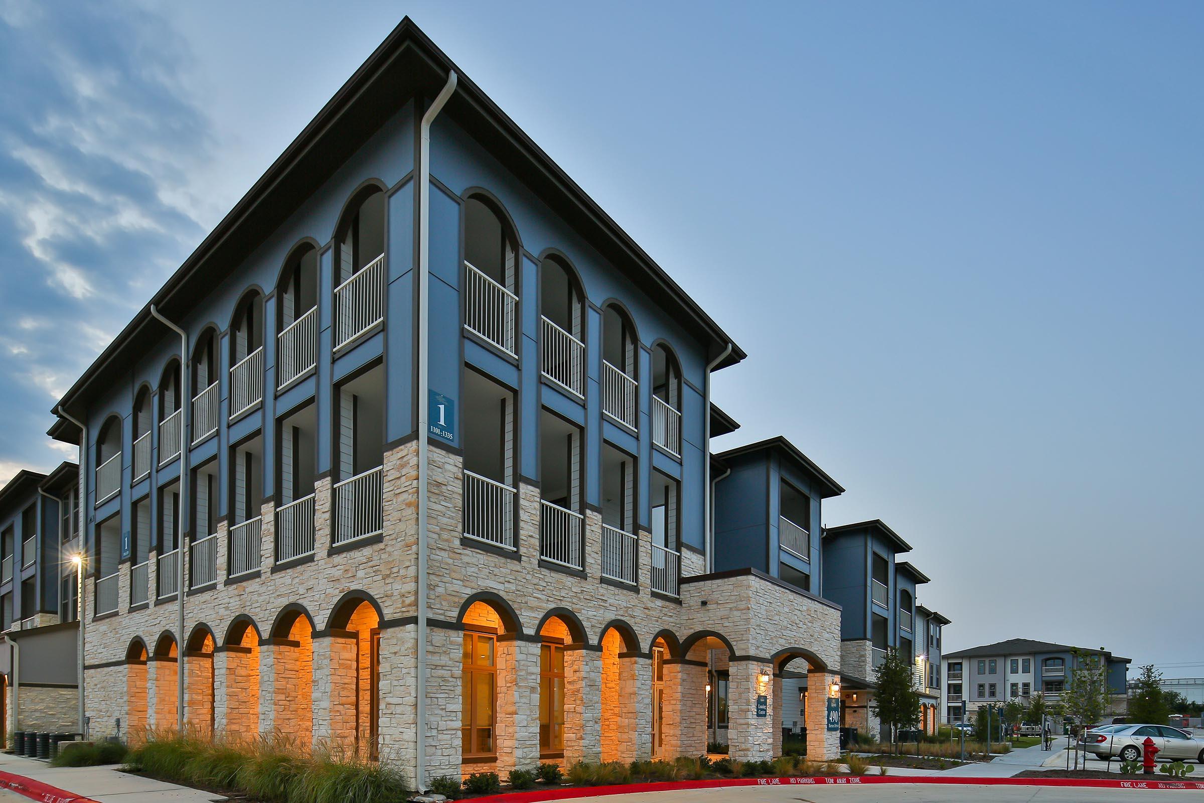 Modern apartment building with a stylish facade featuring dark blue siding and stone accents. The structure has multiple balconies and arched openings. Soft lighting illuminates the entrance, and the surrounding area has well-maintained landscaping, set against a cloudy evening sky.