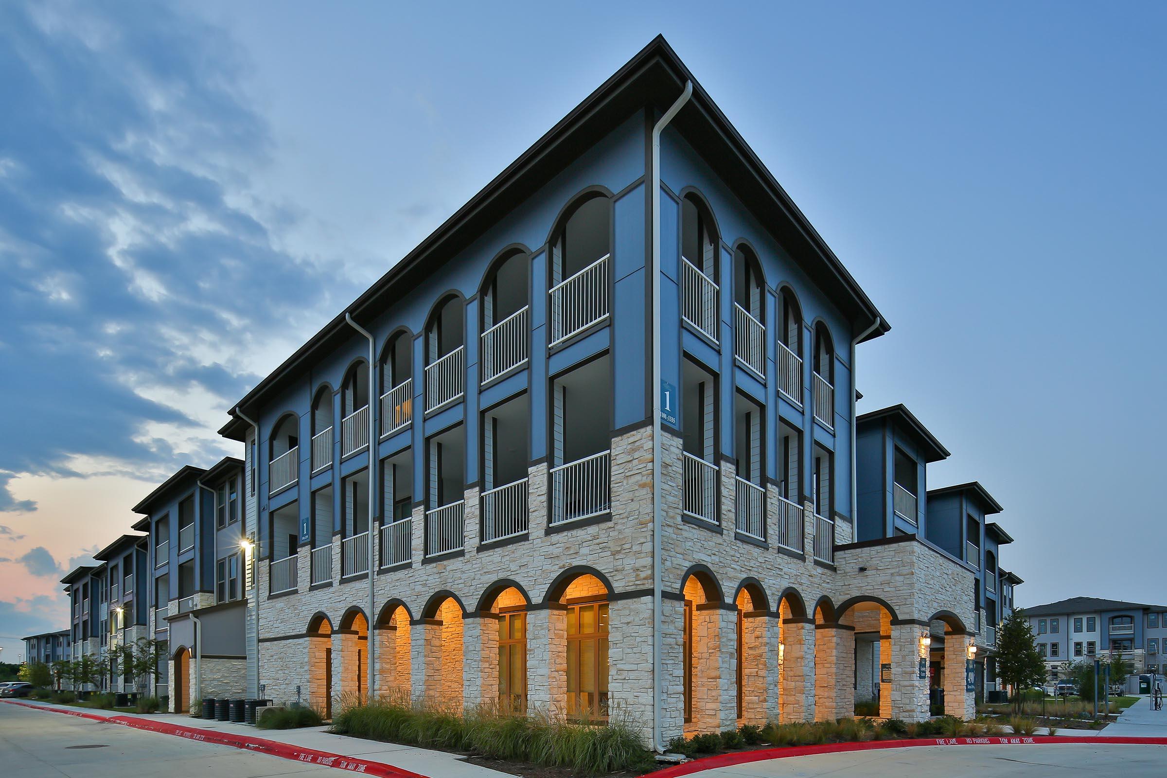 Modern apartment building with a contemporary architectural design, featuring multiple stories, arched windows, and a stone façade. The exterior is illuminated by warm lighting, set against a twilight sky. Surrounding landscaping adds greenery to the scene, enhancing the overall aesthetic.